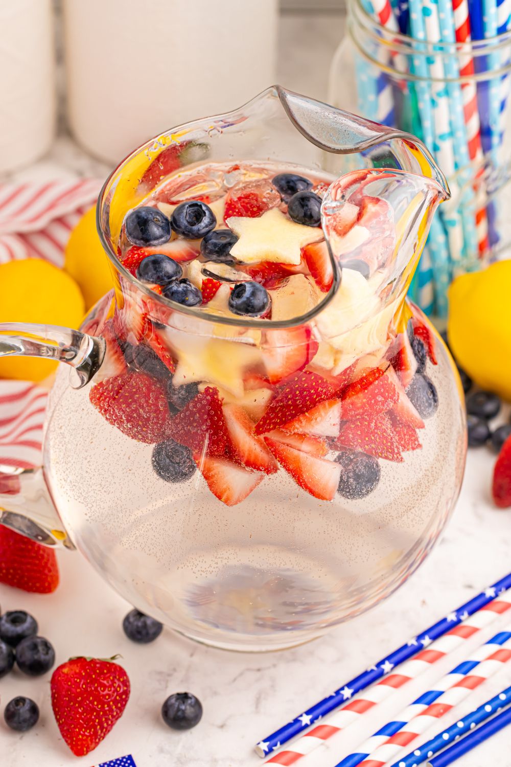 Patriotic Sparkling Lemonade in a container on table with blueberries and strawberries, with paper patriotic straws scattered on table 