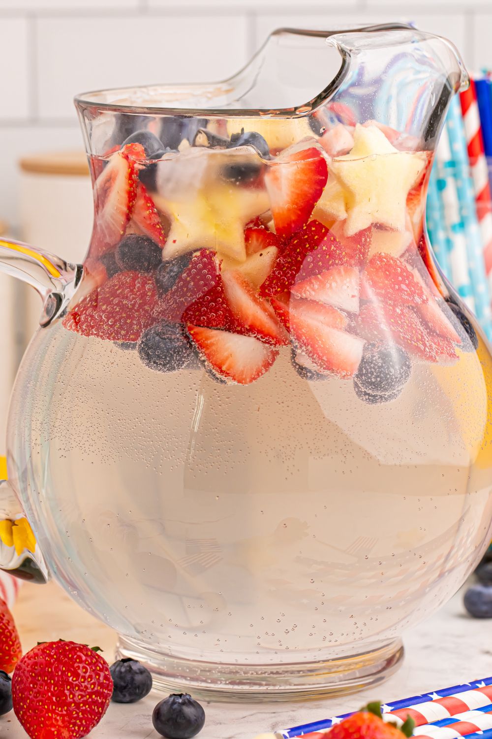side view of glass pitcher with strawberries, blueberries fruit around the table for the Patriotic Sparkling Lemonade