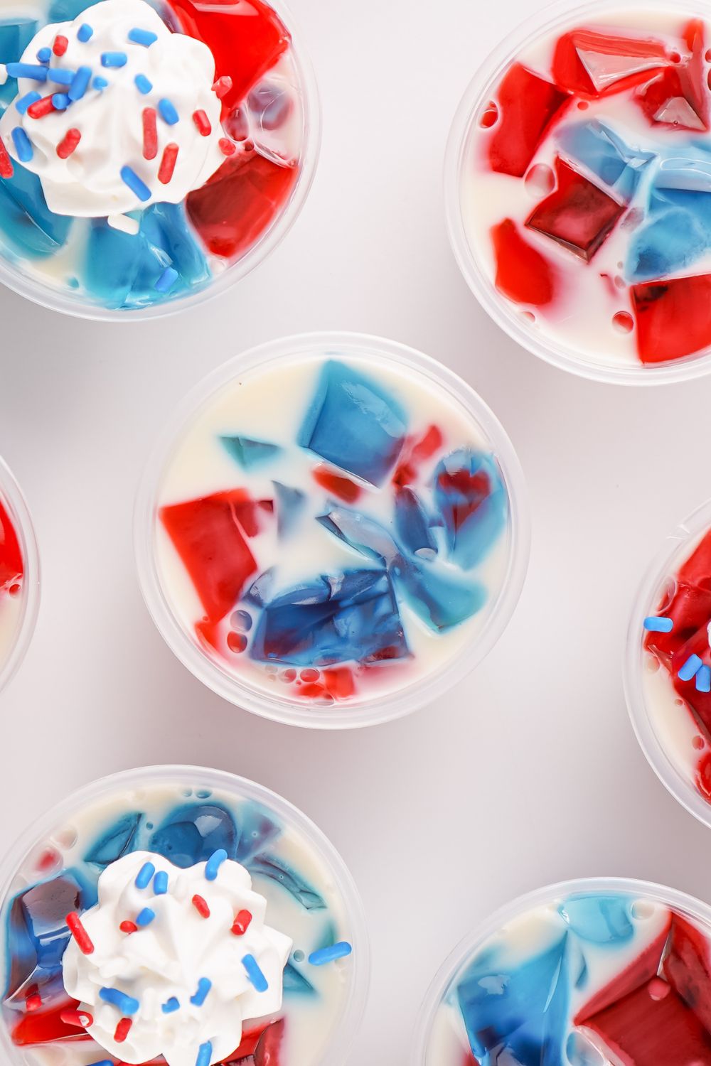 Red White and Blue Jello Shots sitting on table with some having whipped cream and sprinkles on top of them 