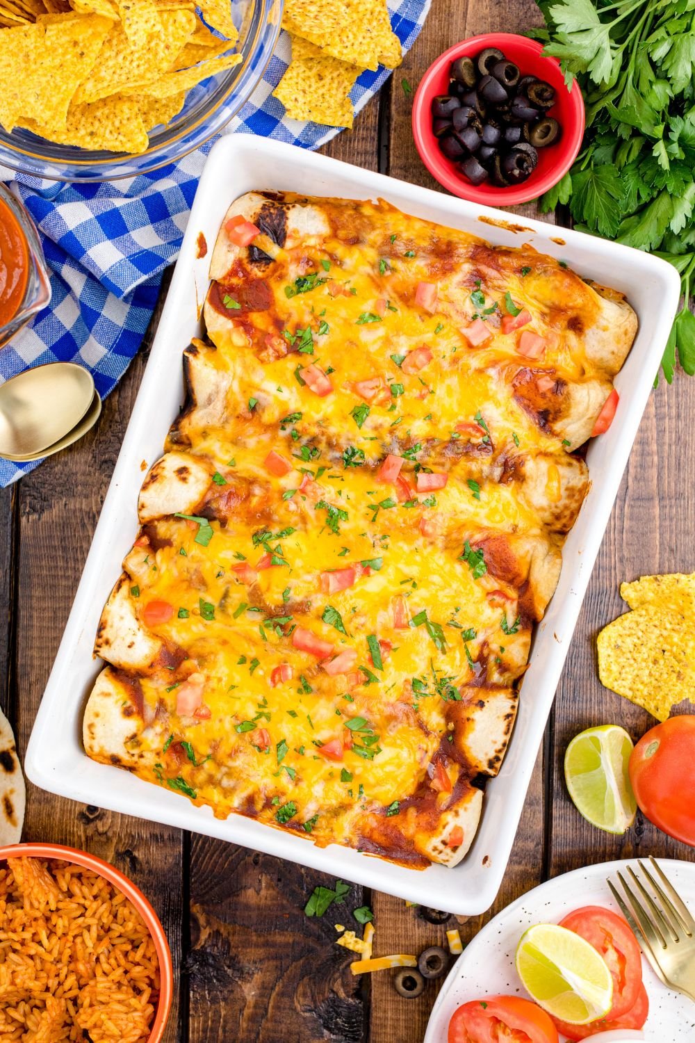 looking down at table to see a casserole dish full of Cheesy Chicken Enchiladas with limes, tomatoes and more around the pan 