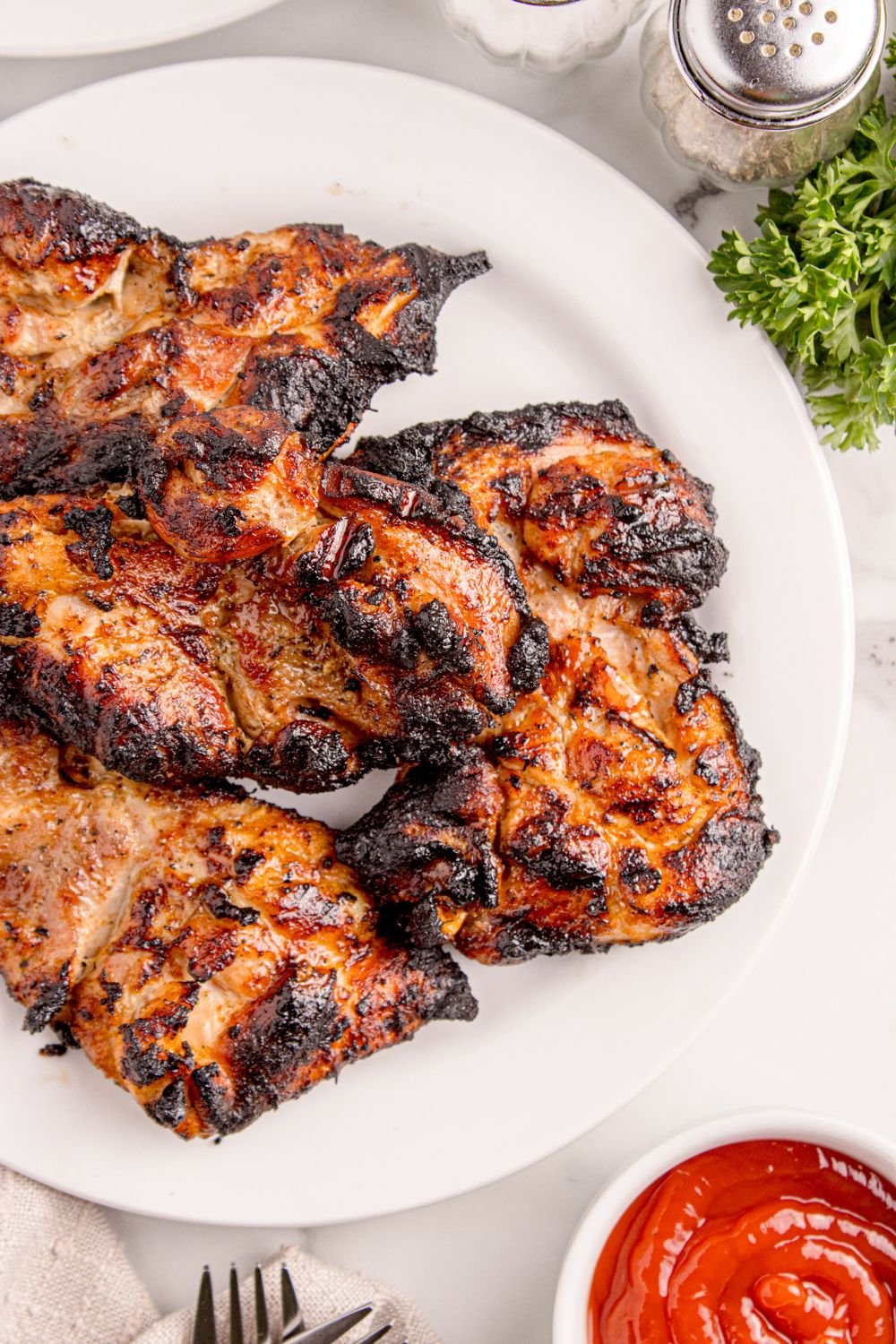 Grilled Chicken Thighs on plate with herbs to side, and a bowl of ketchup in front of plate 