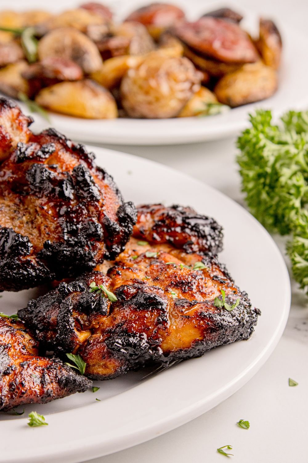 Grilled Chicken Thighs on plate with a plate of cooked potatoes behind it and parsley to the side