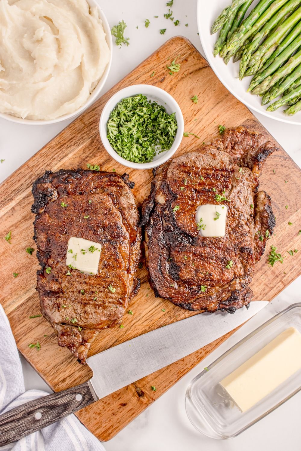 Grilled Ribeye Steaks on a cutting board with slabs of butter and bowl of parsley to the side 