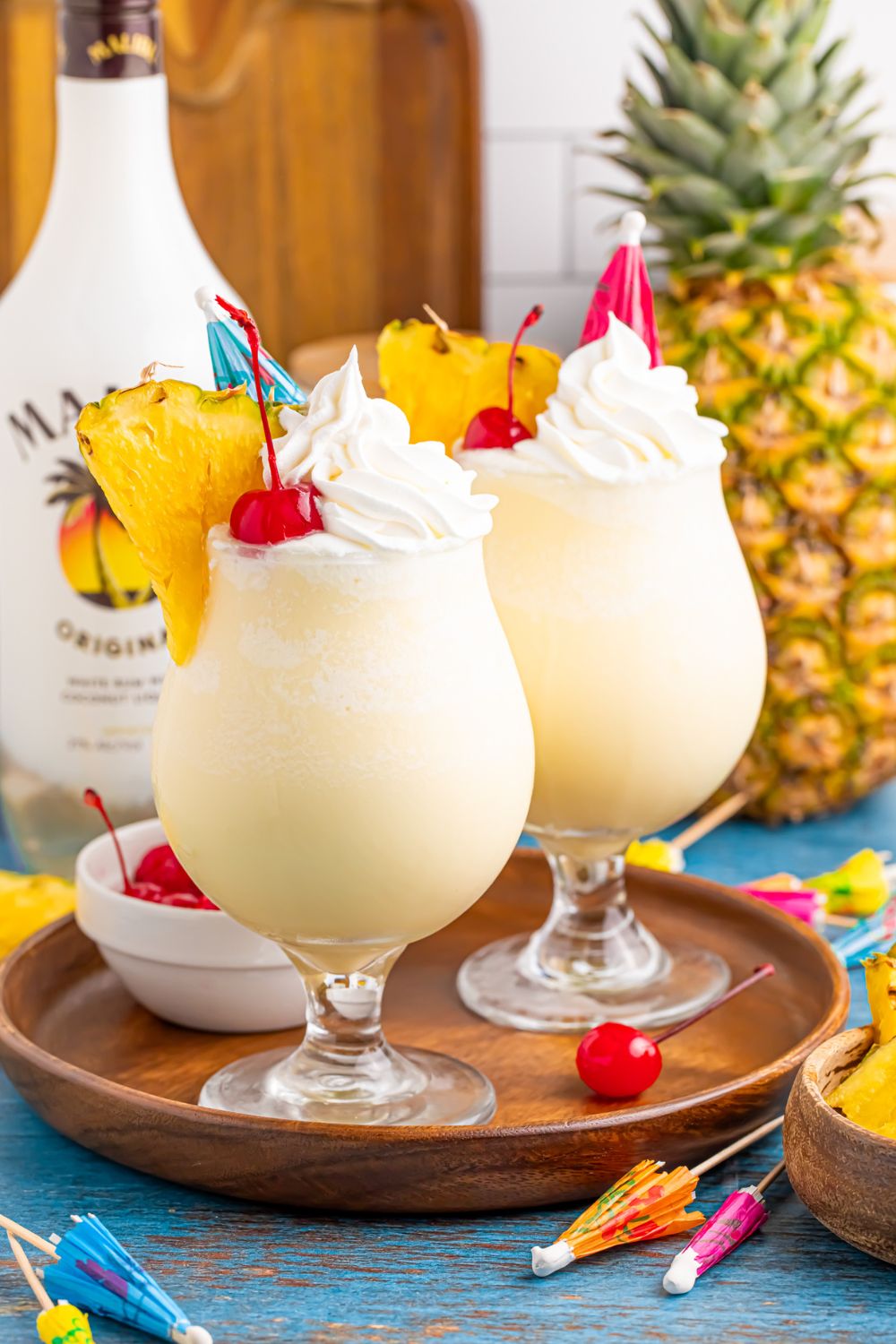 Frozen Malibu Piña Coladas on a tray with cherries and drink umbrellas scattered on table 