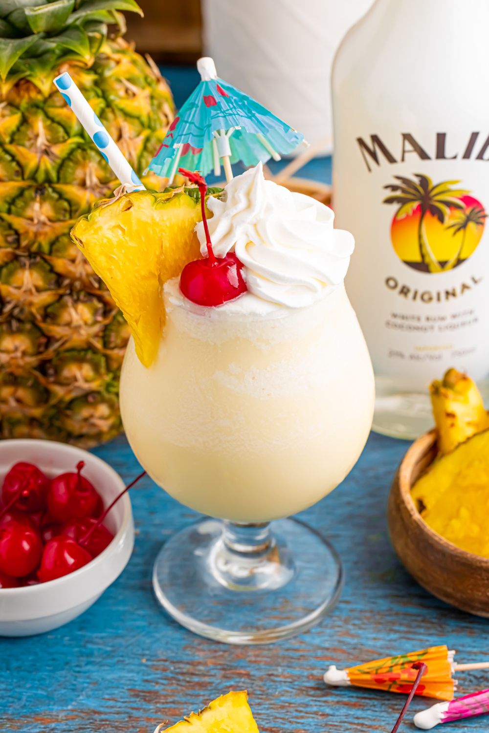 Frozen Malibu Piña Colada in a cocktail glass with a whole pineapple to side, bowl of cherries, and bottle of Malibu rum