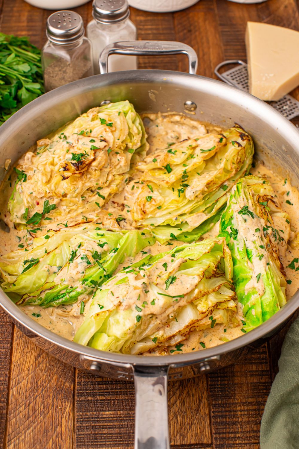 Creamy Garlic Parmesan Cabbage in a skillet sitting on table with a wedge of parmesan to side, salt and pepper, etc