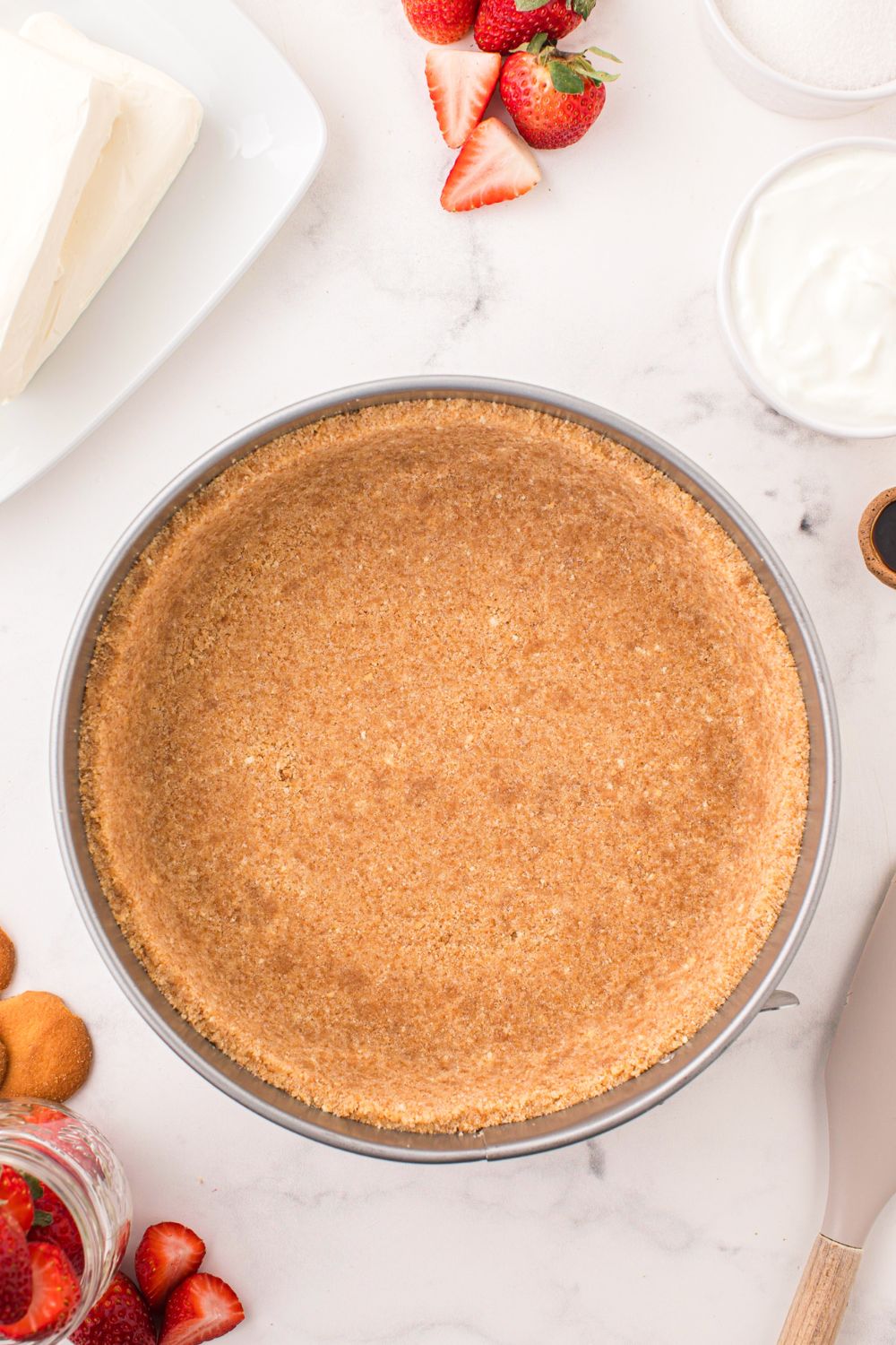 vanilla wafer crust in a pie plate on table with strawberries and other ingredients around it for making a No Bake Strawberry Cheesecake