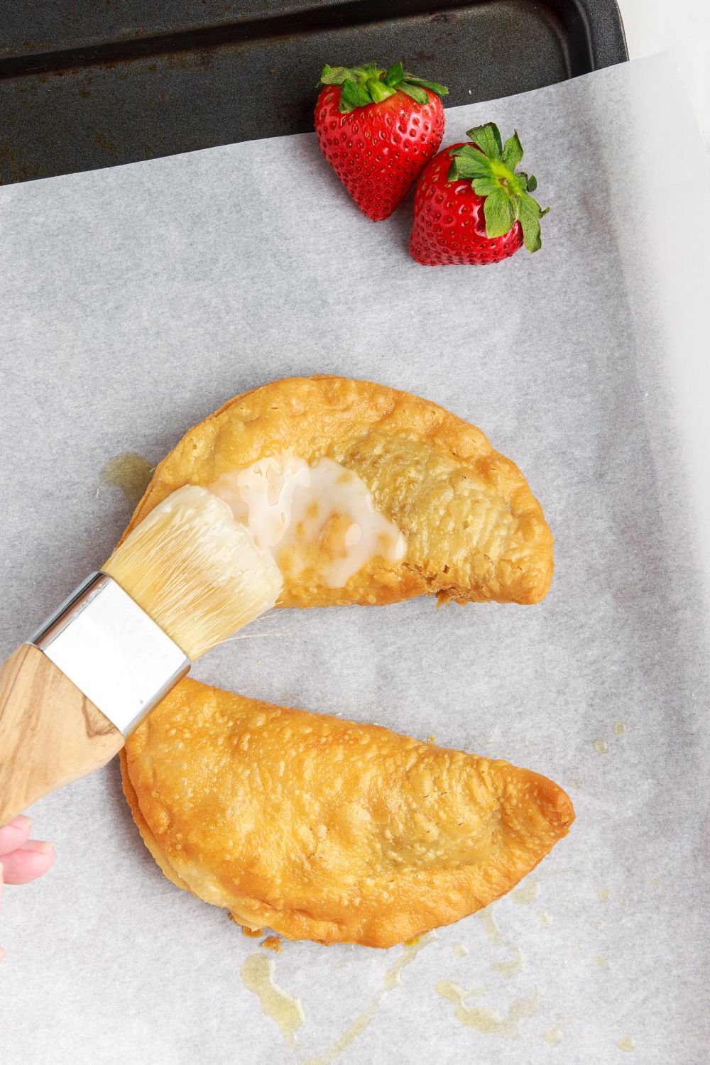 Deep Fried Strawberry Cheesecake Pies on parchment lined tray being brushed with glaze 