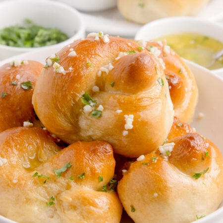 Homemade Garlic Knots piled out on a plate with butter, parsley and dipping sauces to side