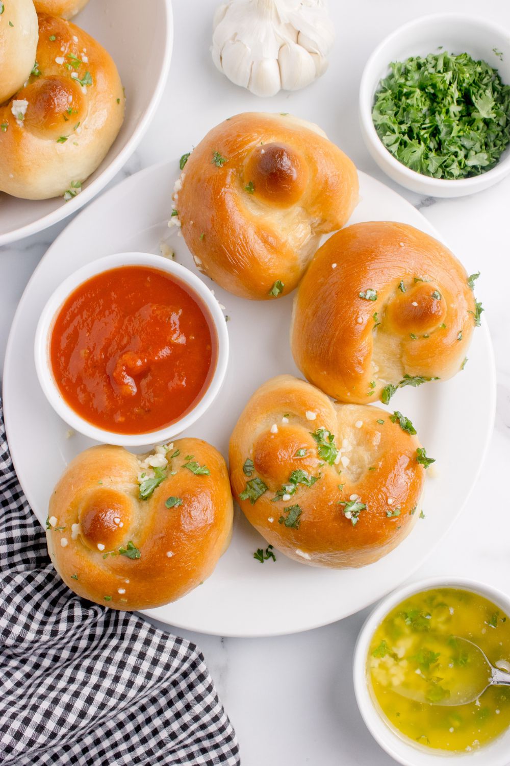 Homemade Garlic Knots spread out on plate with some marinara on it, and a side of garlic butter sitting on table beside the bowl 