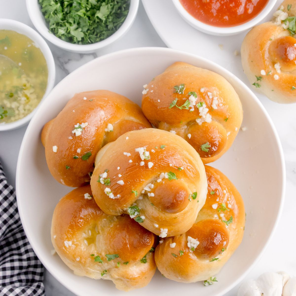 Homemade Garlic Knots piled out on a plate with butter, parsley and dipping sauces to side