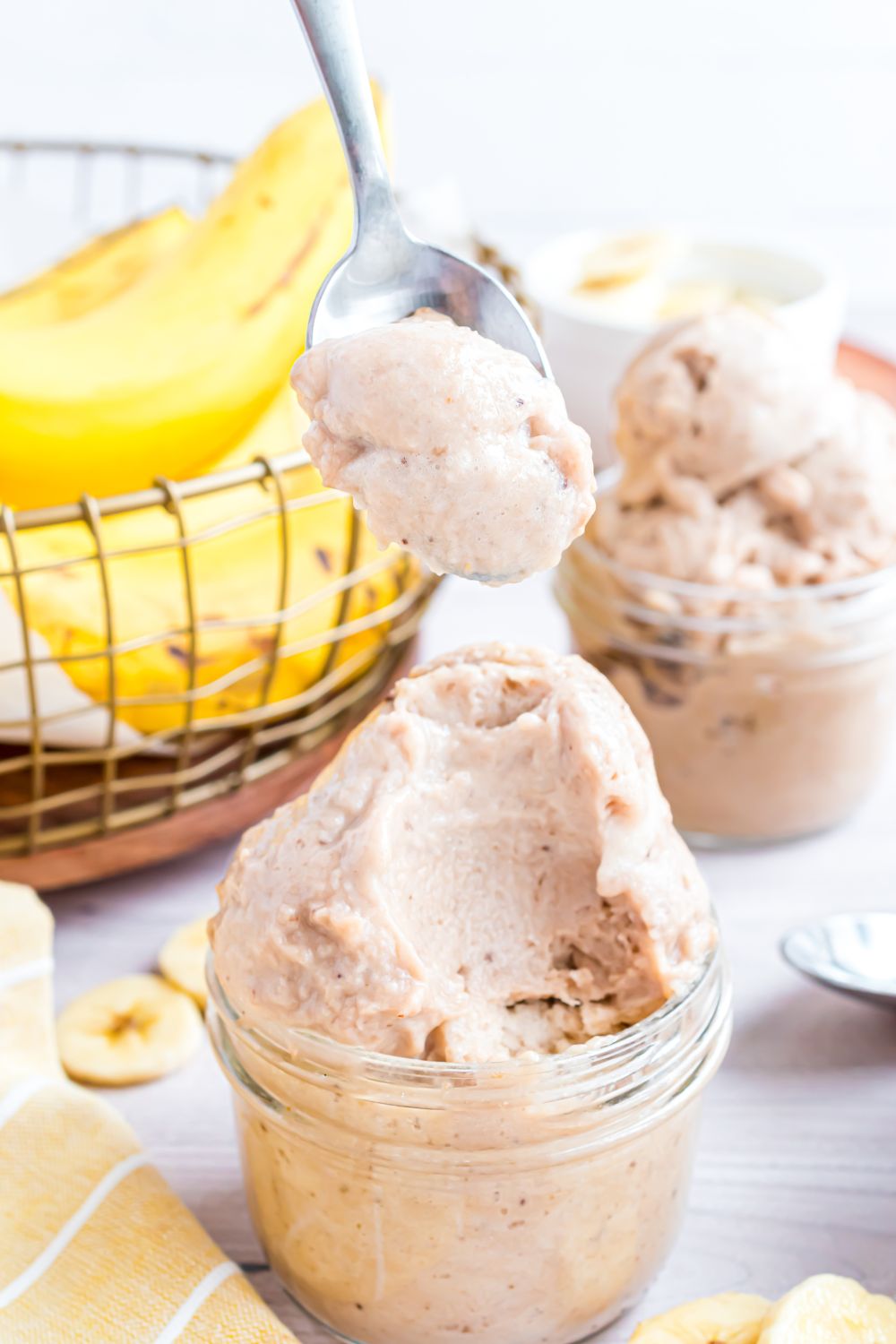 Banana Ice Cream in jars on table with sliced banana to side and a bowl of bananas behind it on table 