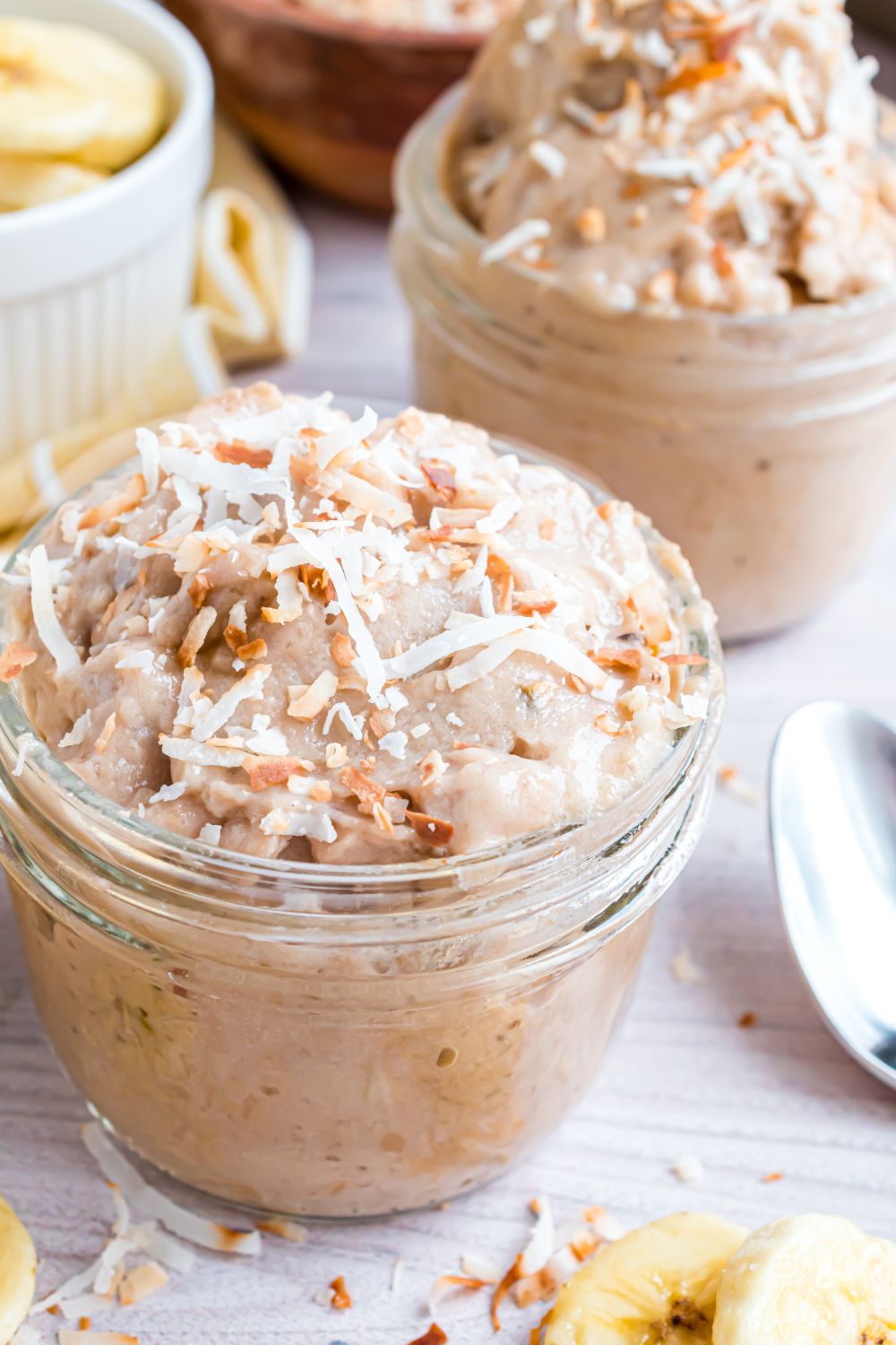 Banana Ice Cream in bowl topped with toasted coconut, and spoons and nuts around table 
