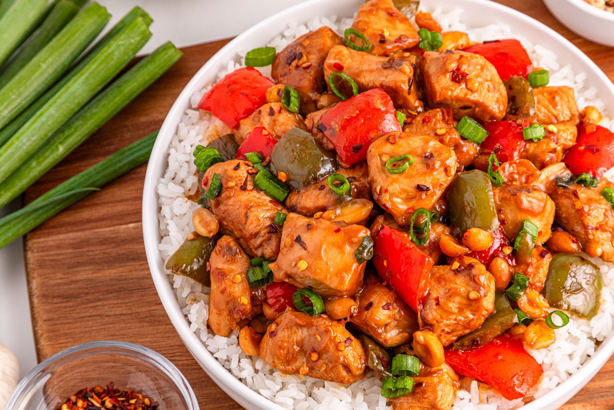 Better Than Takeout Kung Pao Chicken in a bowl with a bed of rice  and cutting board with vegetables to the side 