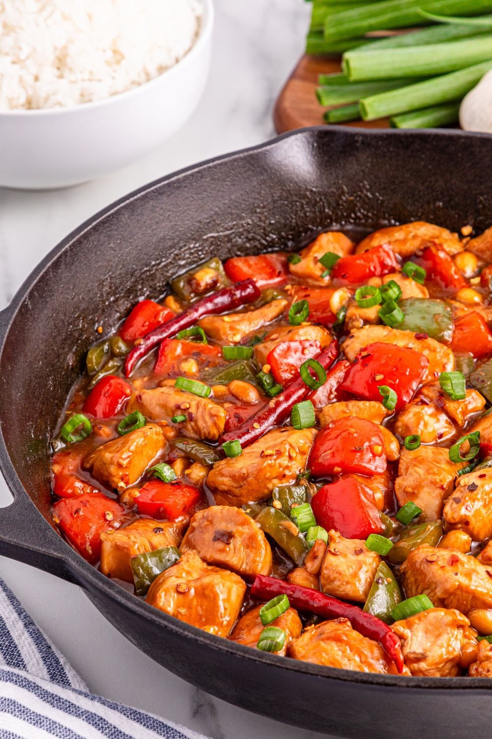 Better Than Takeout Kung Pao Chicken in a cast iron skillet with a bowl of rice to the side and green onions on cutting board 