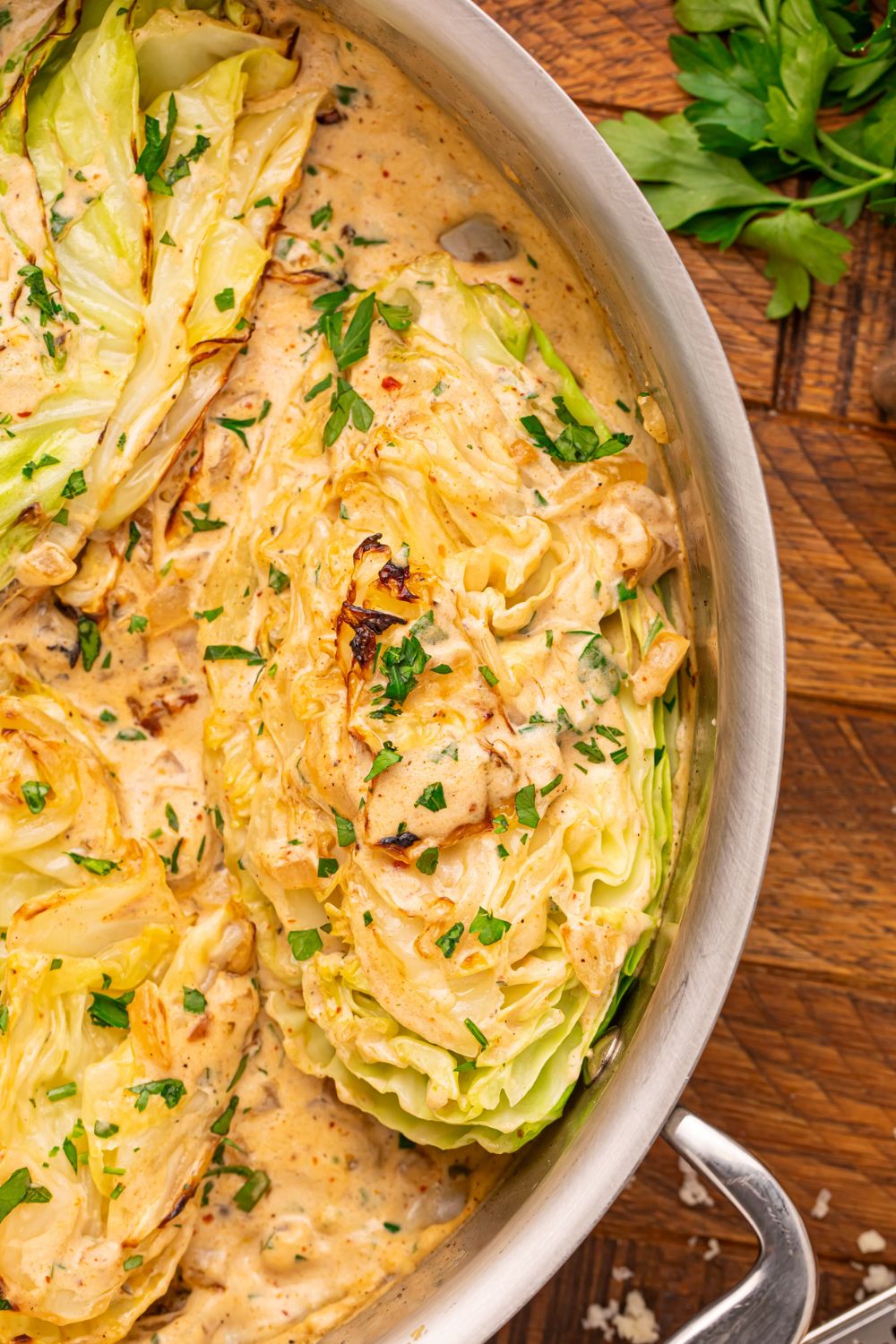 Creamy Garlic Parmesan Cabbage in a skillet sitting on table with a bundle of parsley to side and sprinkled on top of cabbage and sauce in skillet 