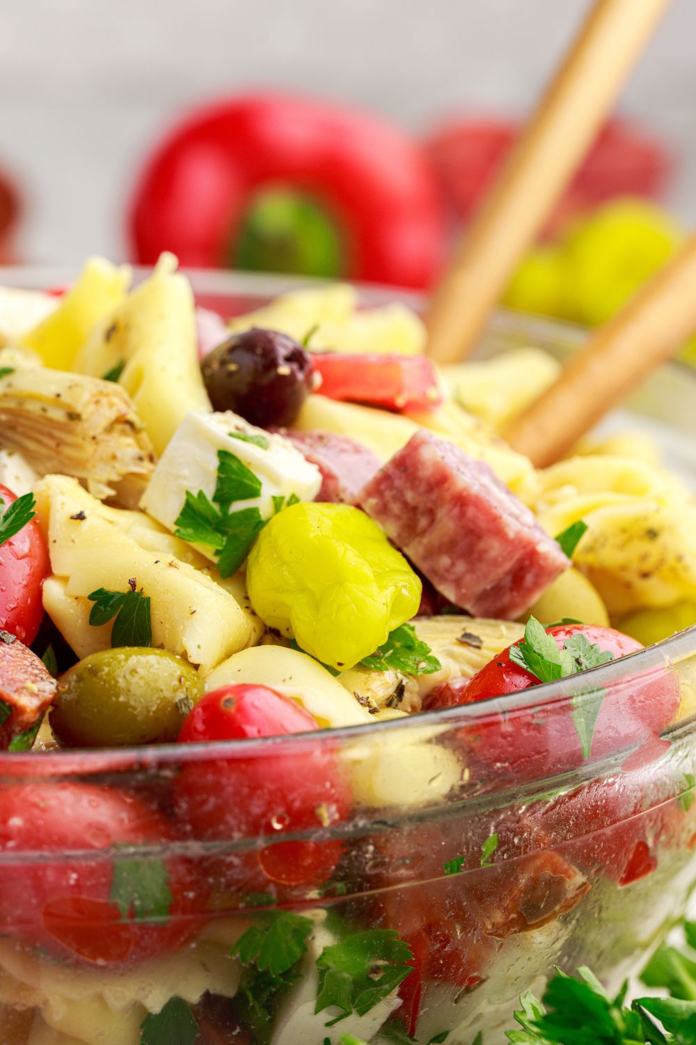 side view of Antipasto Tortellini Pasta Salad in a glass bowl with spoons pushed into bowl and vegetables around the table 