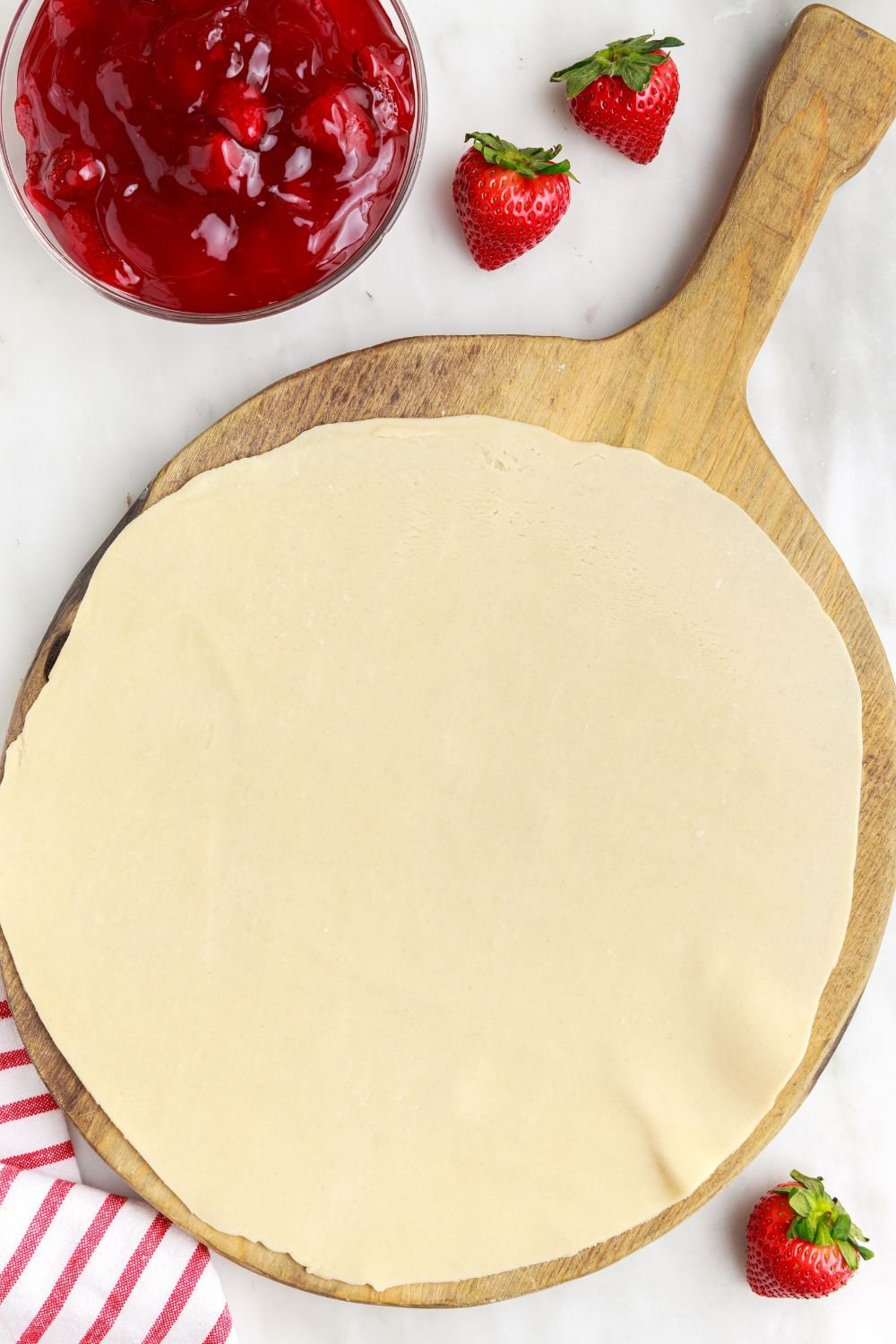 pie crust spread out on a cutting board sitting on table and strawberry pie filling to the side