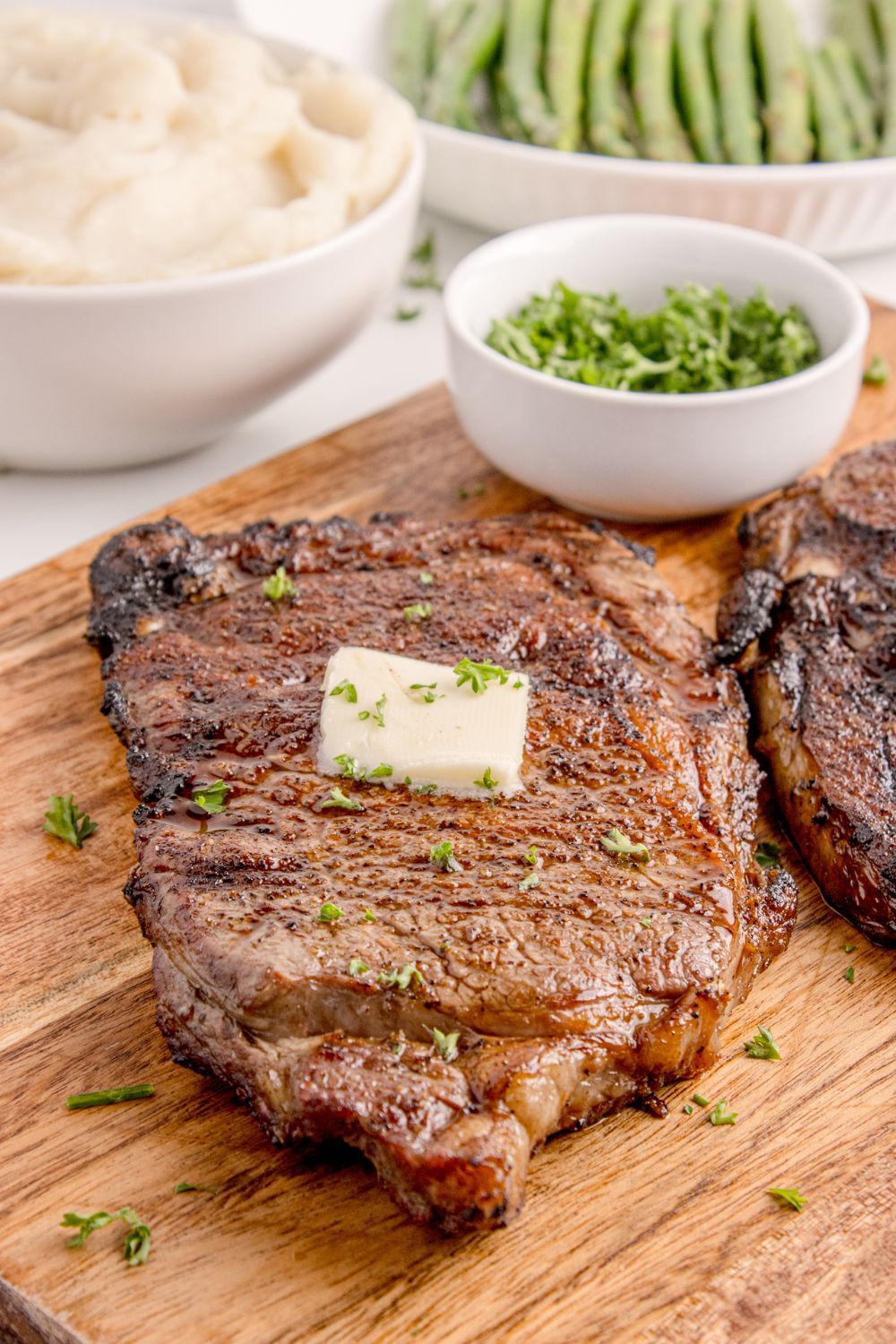 Grilled Ribeye Steak on cutting board with a slab of butter on top of steak and a bowl of parsley to side 