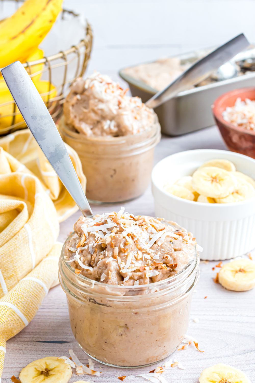 Banana Ice Cream in bowls with toppings on top and spoons in bowls, and bananas sliced in a white bowl on table 