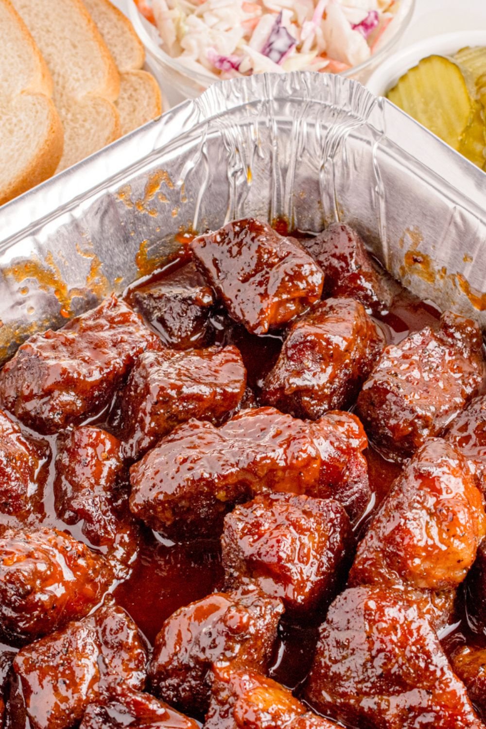 Chuck Roast Burnt Ends in a pan on table with sliced bread to the side and a small bowl of pickles 