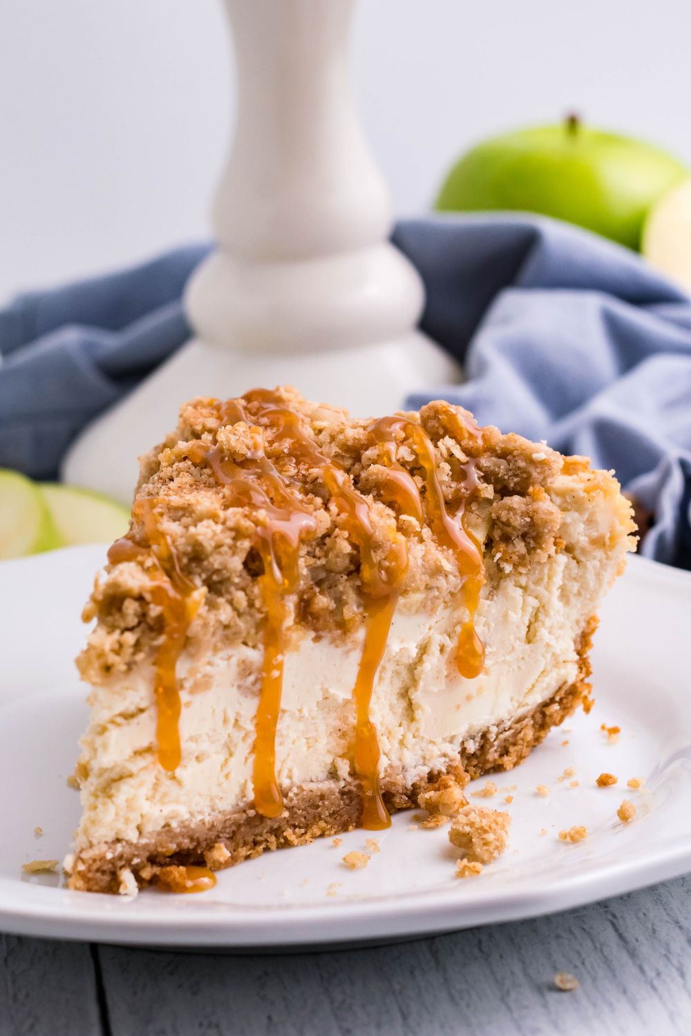 slice of Caramel Apple Crisp Cheesecake on plate with a pie plate behind it on table, napkin and green apples 