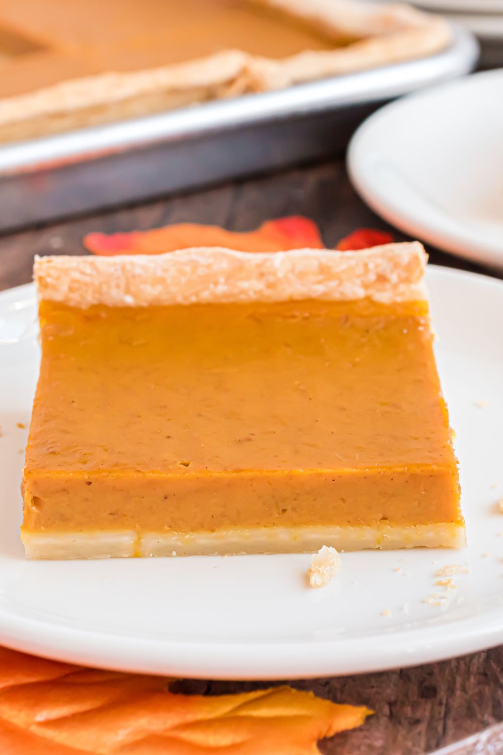 white plate on table with a square slice of pumpkin pie with more plates with pie around table and a Sheet Pan Pumpkin Pie in background on wooden table 