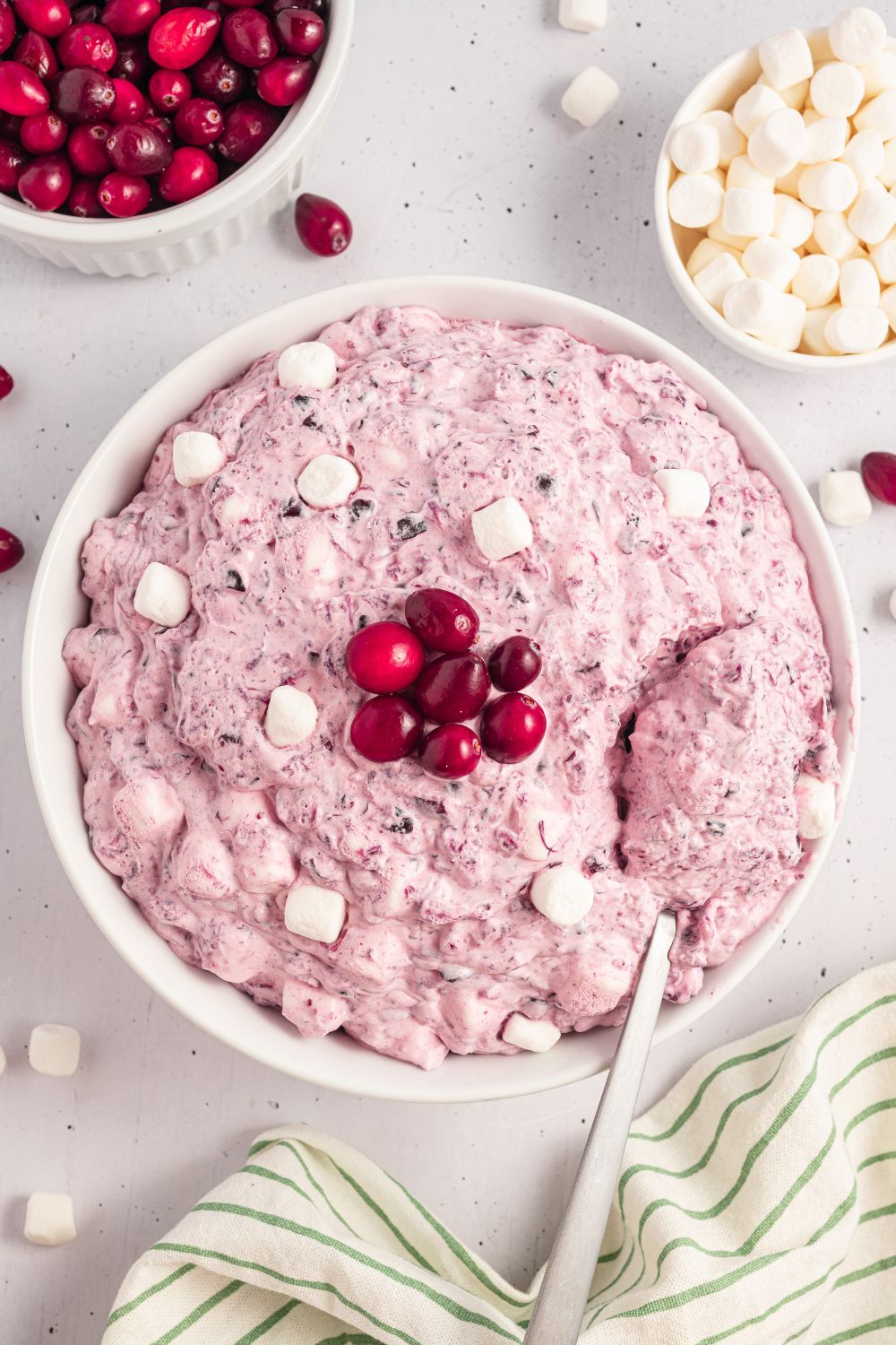 white bowl on table with some Cranberry Fluff Salad topped with extra marshmallows, cranberries, and a mini bowls of cranberries and marshmallows. Then more mini marshmallows on table with a pin striped towel to side 