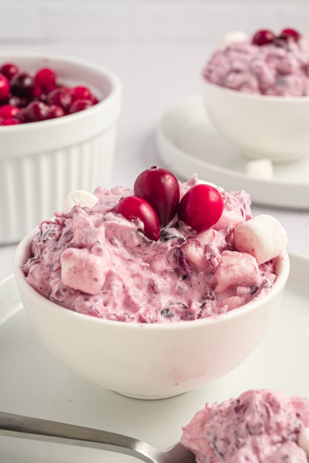white table that has plates with small bowls on top of plate full of Cranberry Fluff Salad topped with a few fresh berries. Then in a bowl in background is more cranberries. 