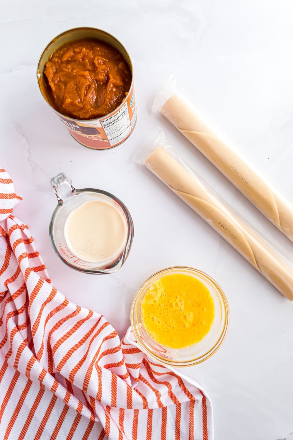 Sheet Pan Pumpkin Pie ingredients on a marble counter. A can of pumpkin puree, two rolls of pie crust, a bowl of egg mixture, and evaporated milk in a measuring cup