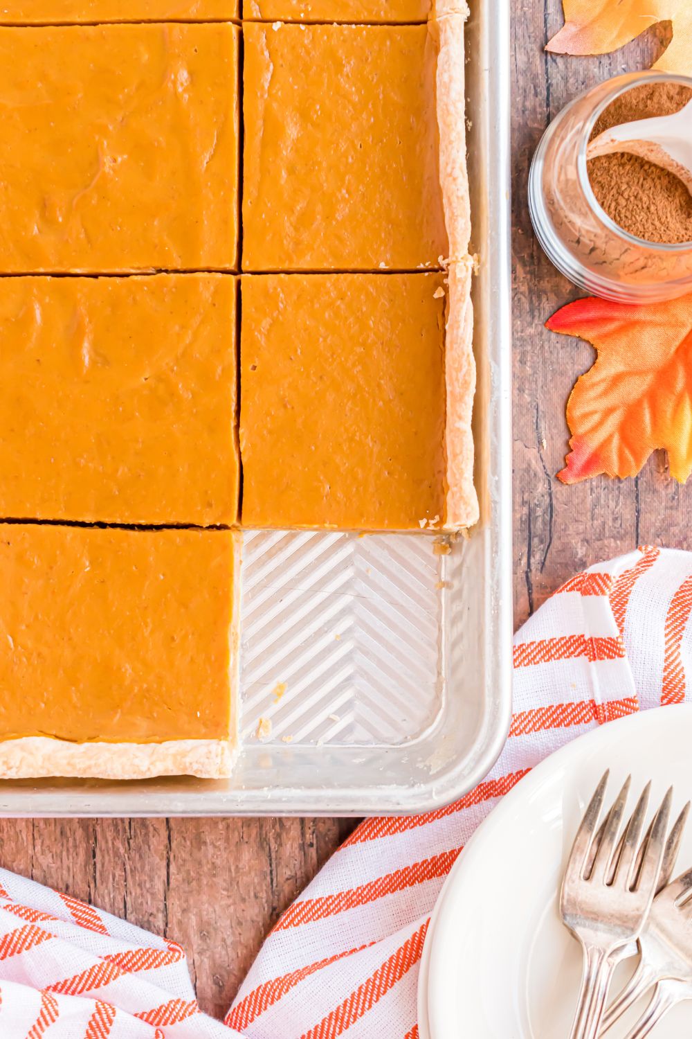 wooden table with a red striped towel spread over the table and behind it is a sheet pan full of baked pumpkin pie with one slice removed from sheet pan decorated for Thanksgiving 