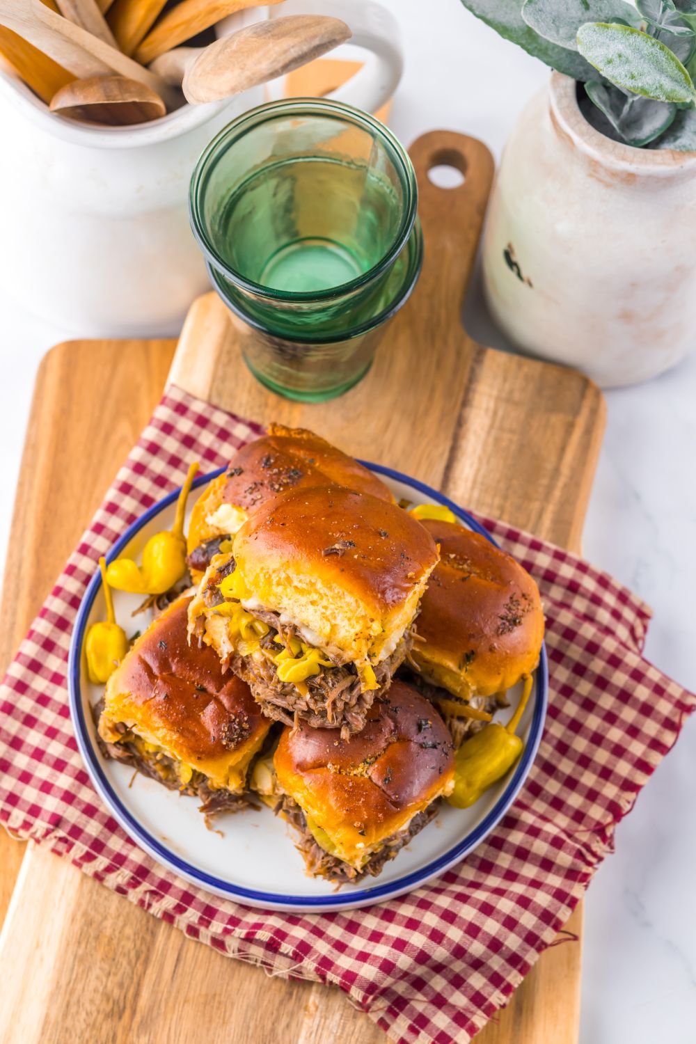 Slow Cooker Mississippi Pot Roast Sliders stacked on a plate with some glasses and ceramic containers behind it on table 