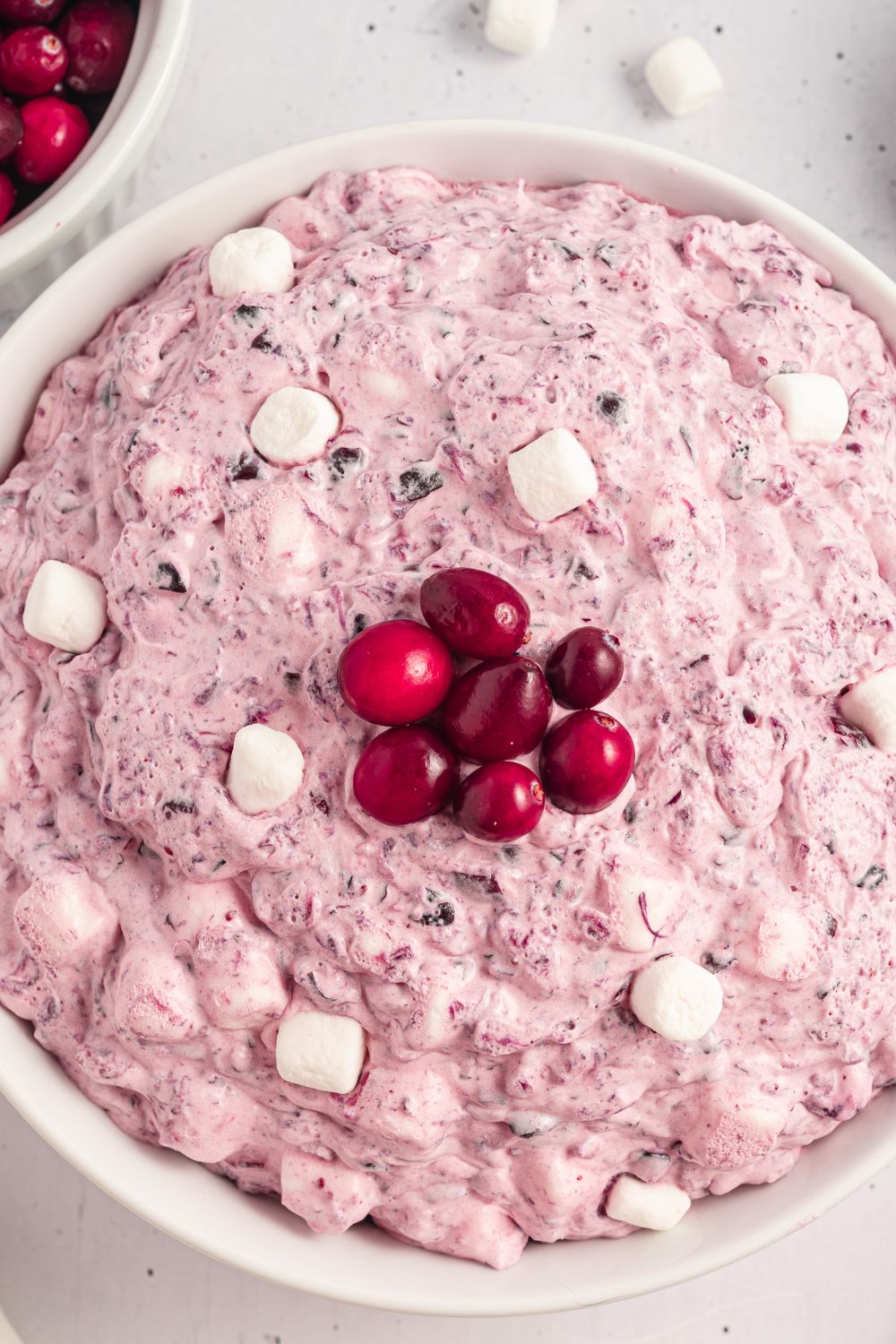 view of a white bowl on table with marshmallows and cranberries with a few mini marshmallows scattered around table 
