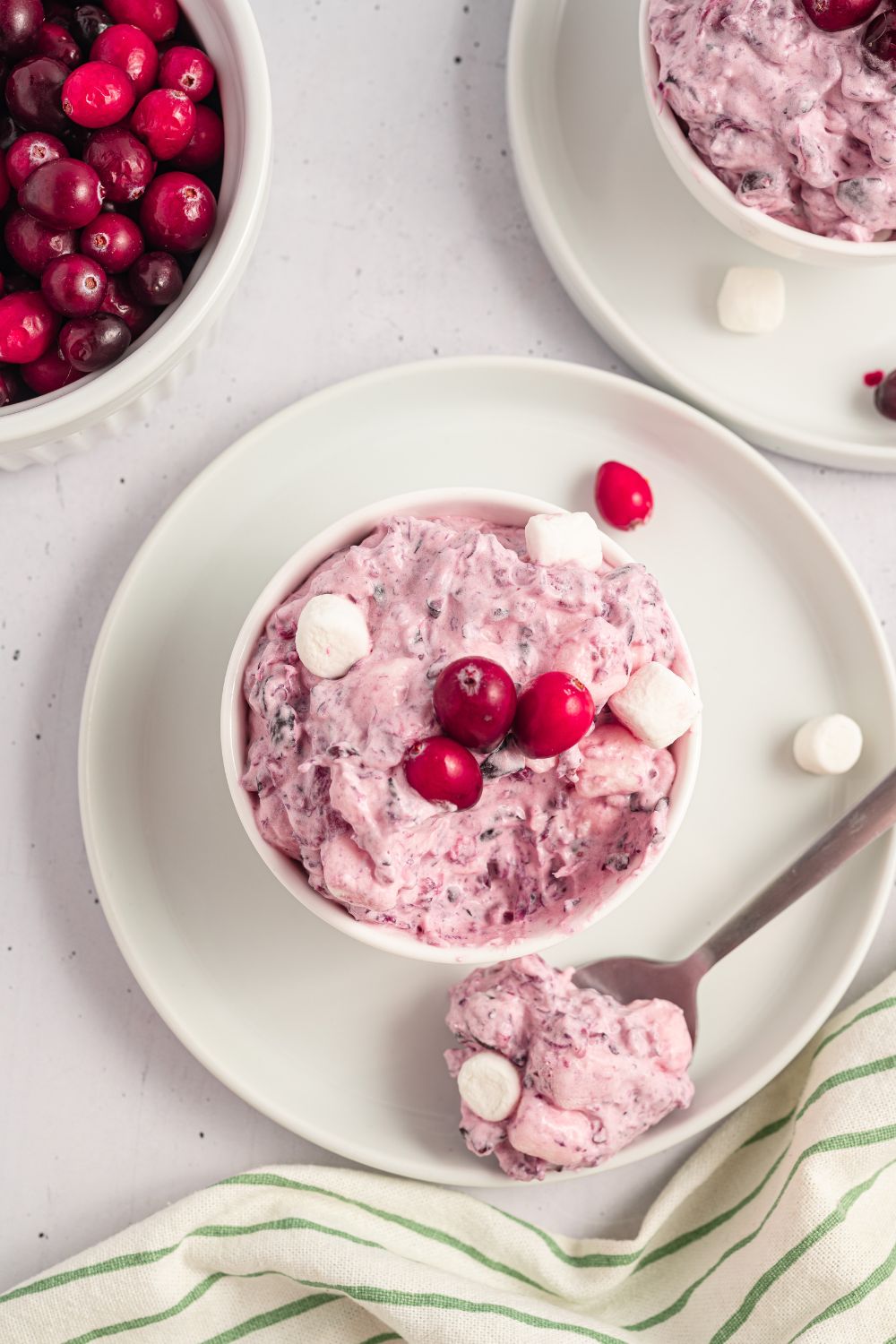 white table with a plate with a small bowl full of Cranberry Fluff Salad, and a spoonful of salad sitting on edge of plate. A bowl of fresh cranberries to the side of the plate 