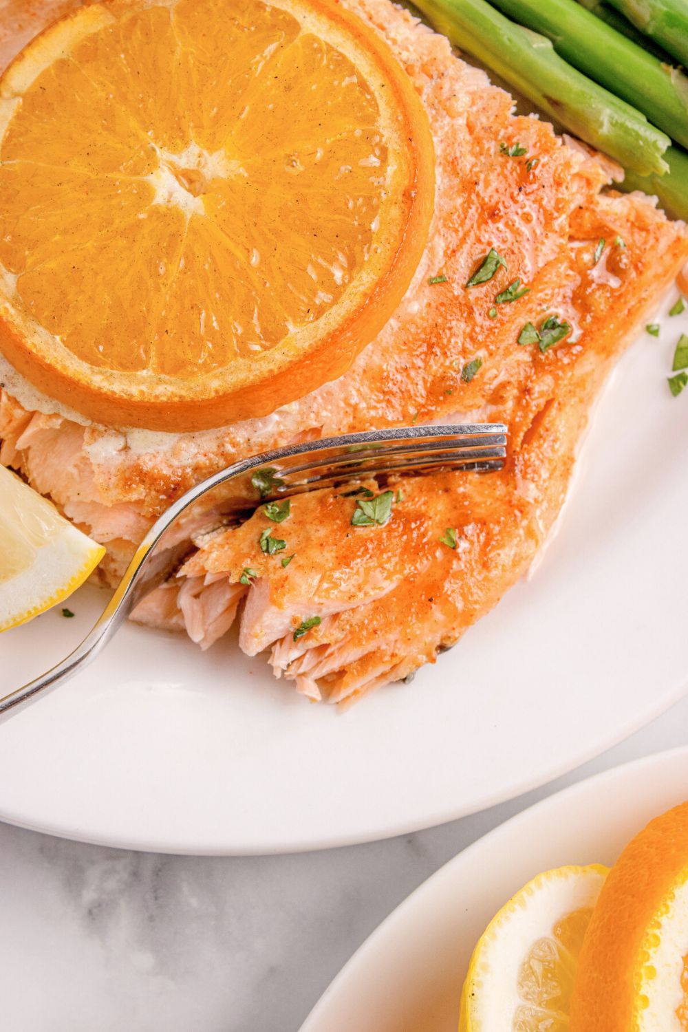 salmon on plate with an orange on top, with a fork cutting into the salmon, and asparagus to side