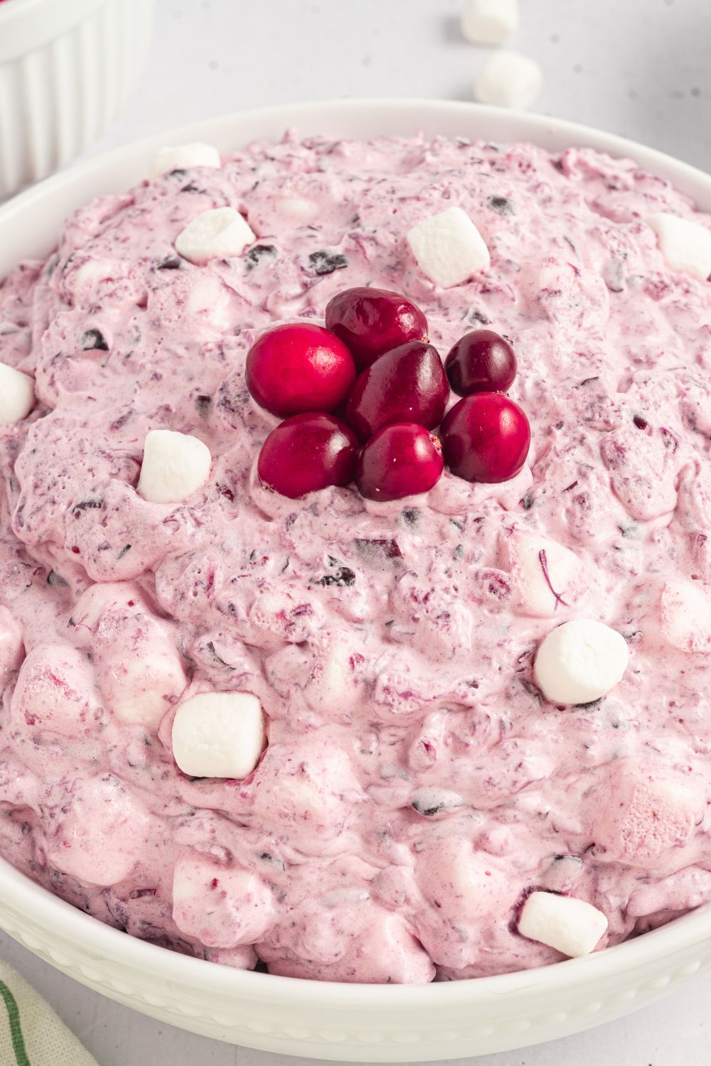close up of a white bowl full of Cranberry Fluff Salad with mini marshmallows around the top of fluff, and in the center there are 7 fresh cranberries in the center of bowl 