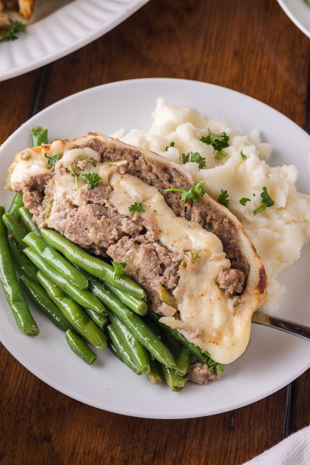 wooden table with a white plate sitting on it, that has green beans, philly cheesesteak meatloaf, and a spoonful of mashed potatoes