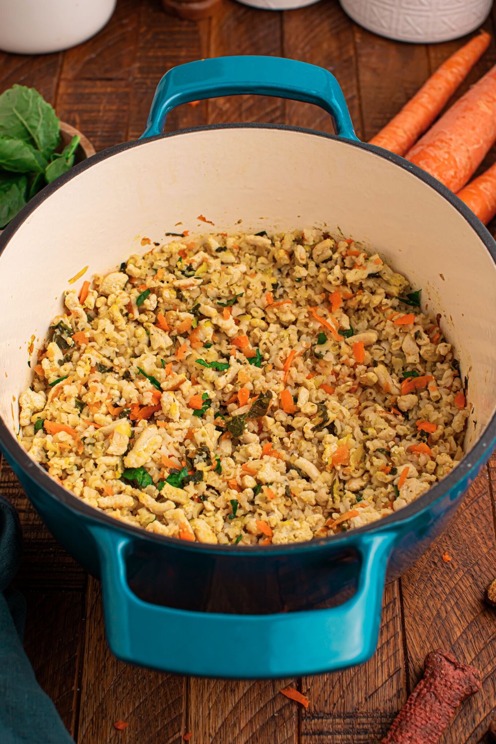 Homemade Turkey and Rice Dog Food with Veggies in a pot sitting on table with fresh herbs, carrots, and white containers behind pan on table 