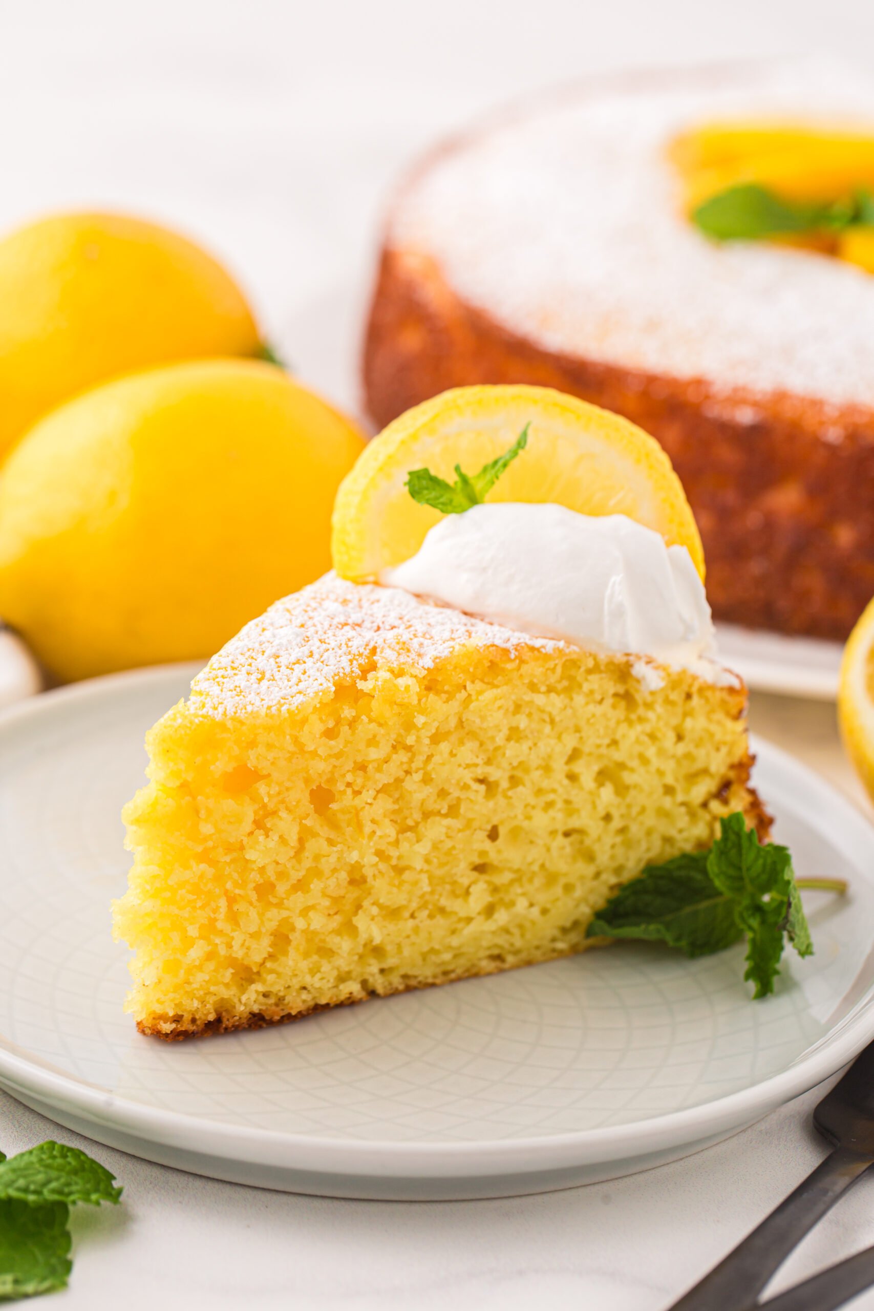 slice of Italian Lemon Ricotta Cake on a white plate with whole lemons behind it on table and the rest of the cake behind it
