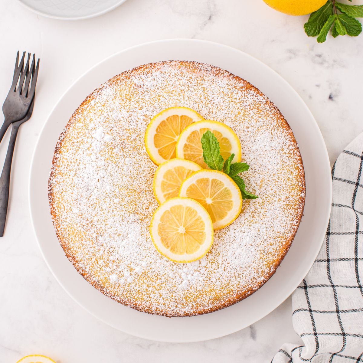 Italian Lemon Ricotta Cake on table with some fresh lemons on top with some mint leaves to side 