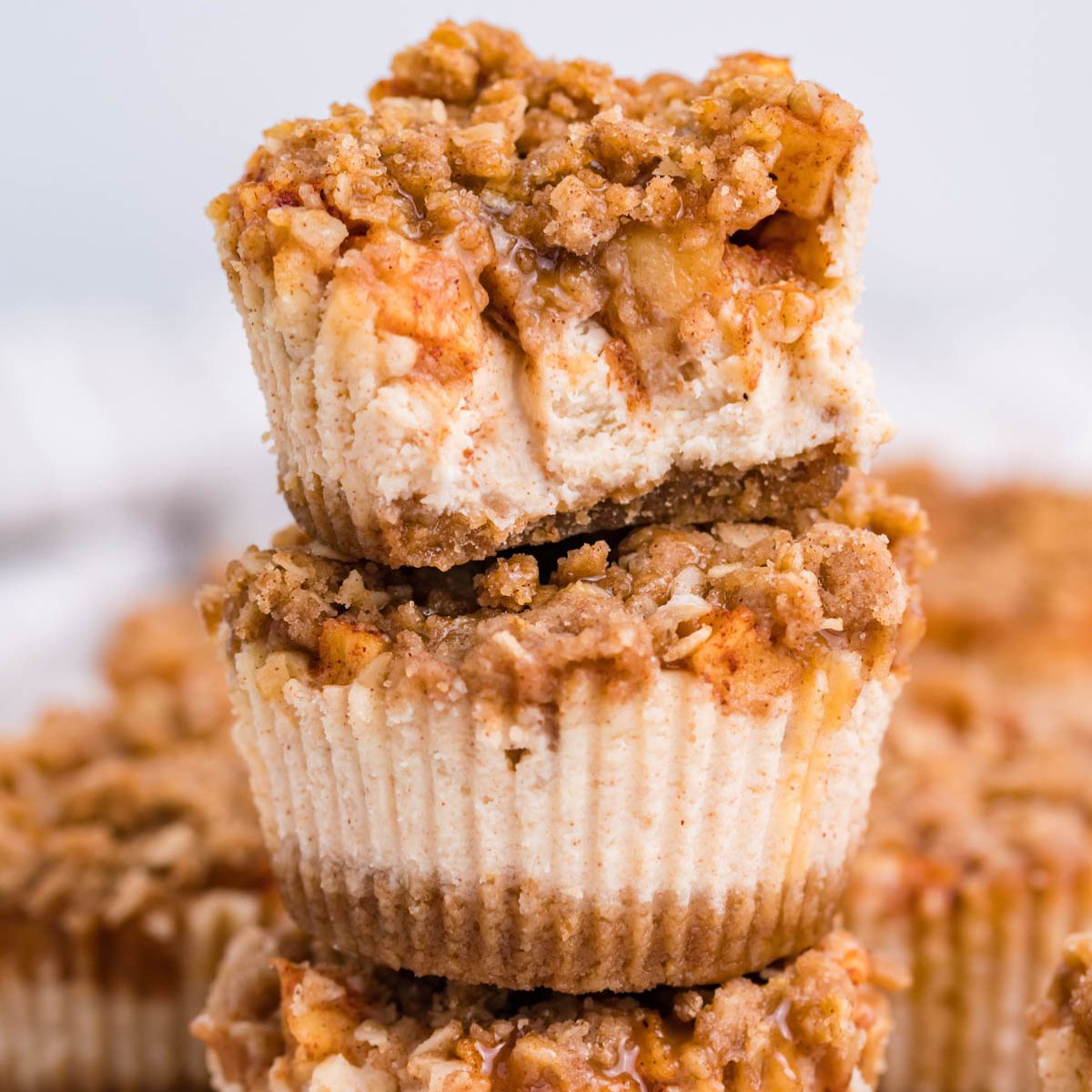 Mini Apple Crisp Cheesecakes stacked on top of one another on table with more spread out behind it on table 