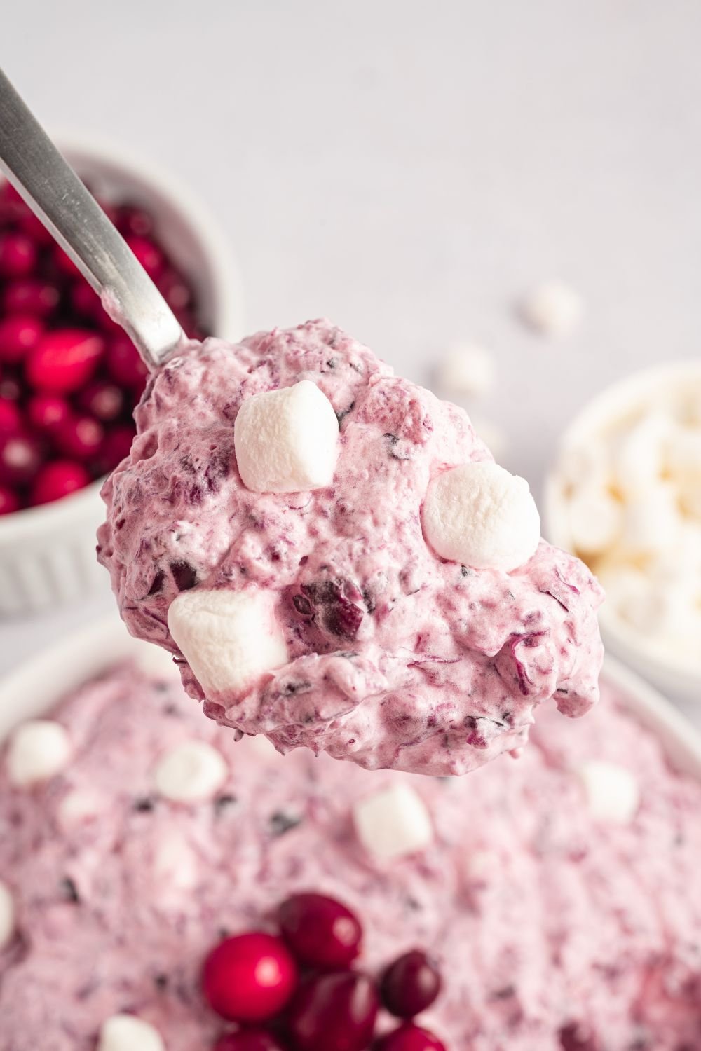 Cranberry Fluff Salad in a white bowl on table with a spoonful of the mixture. Small bowls to side on white counter with marshmallows and cranberries 