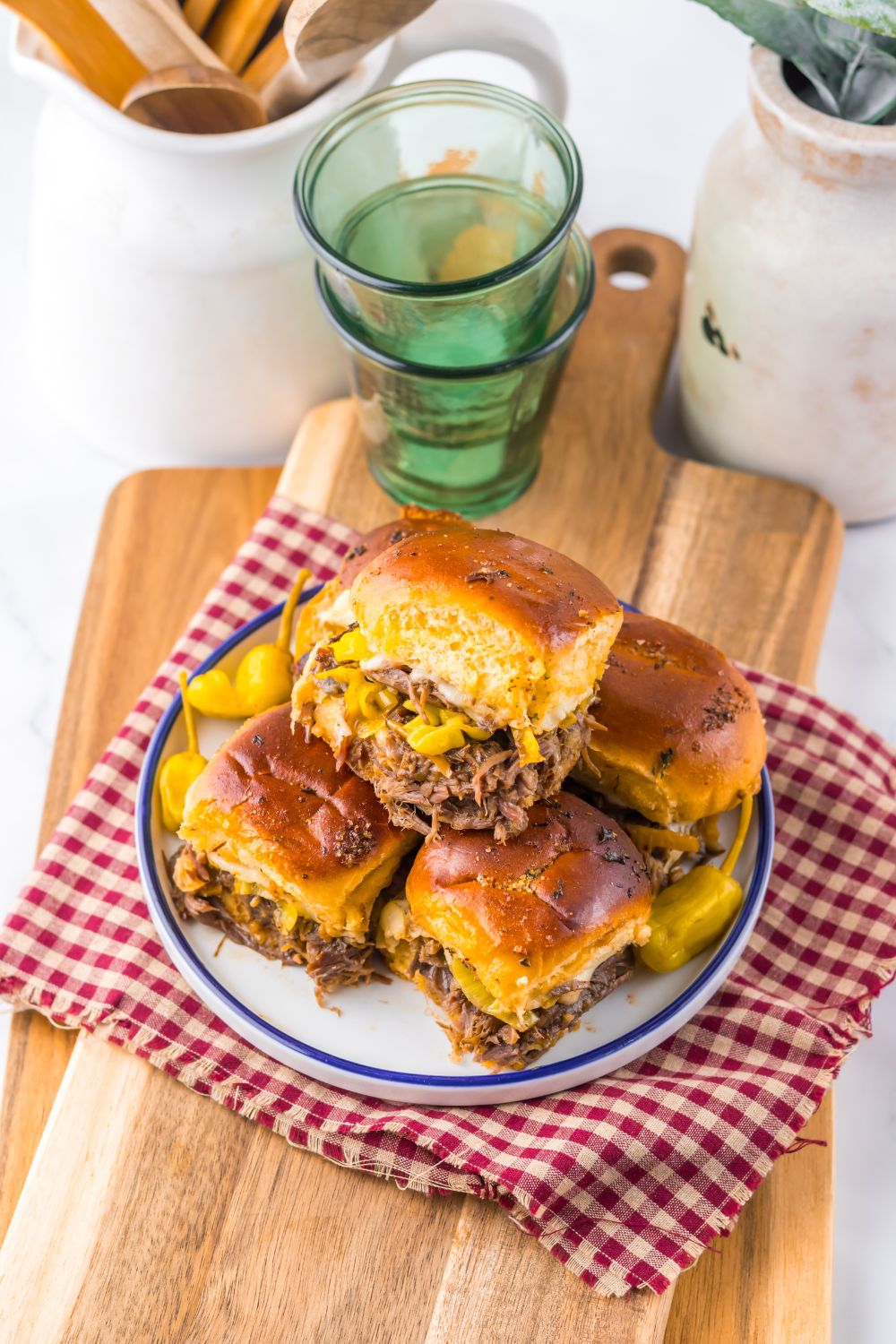Slow Cooker Mississippi Pot Roast Sliders stacked on a plate with a towel under it with some glasses behind it on table 