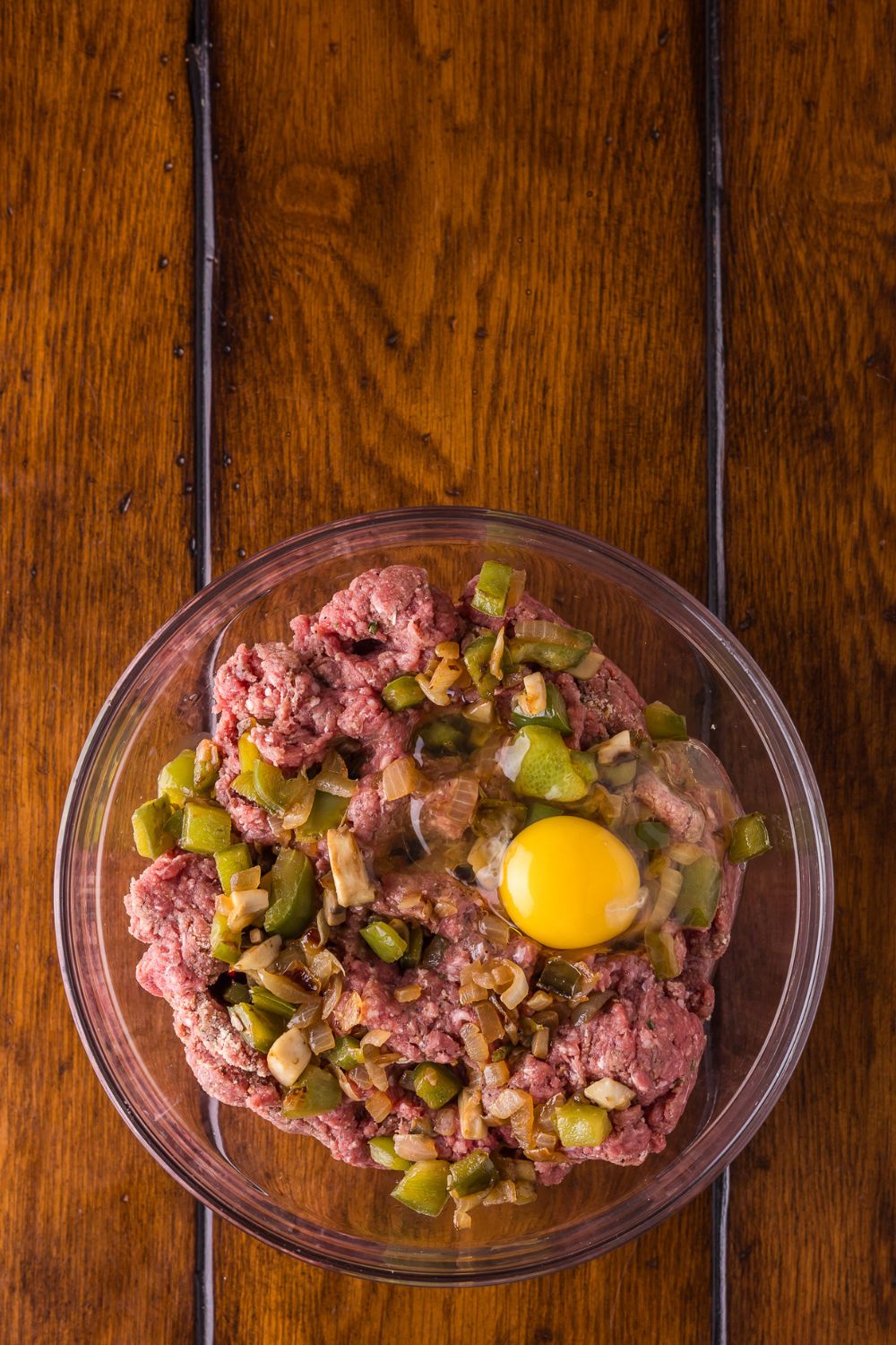 raw meat, with egg, and onions and peppers on a glass bowl on a wooden table