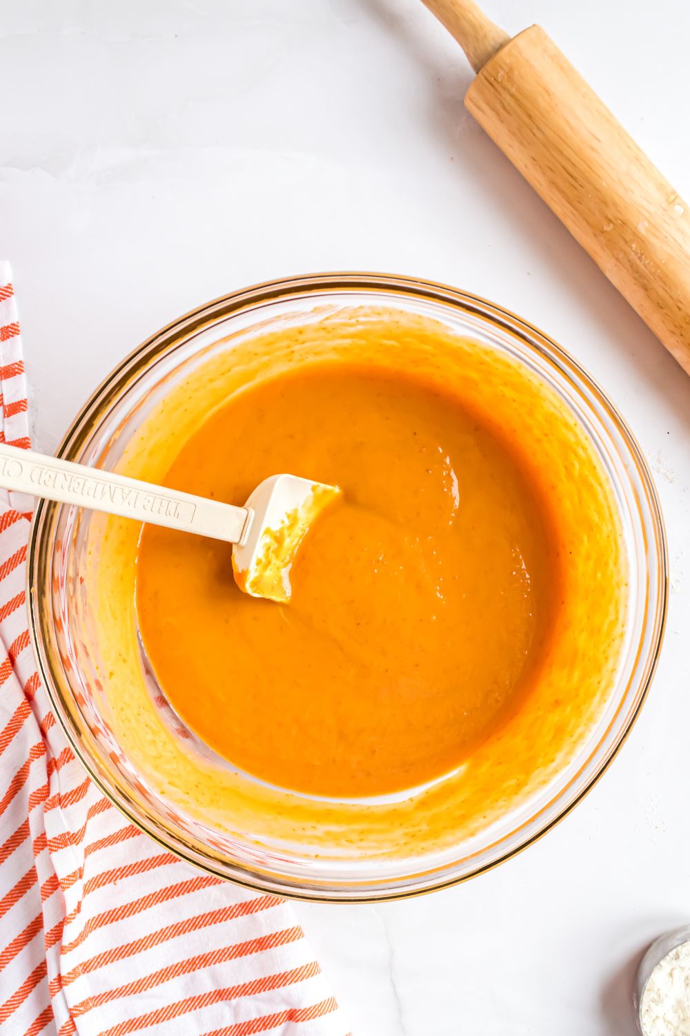 glass bowl sitting on white table that has pumpkin pie batter fully mixed up with a red striped towel to side and rolling pin to the side 