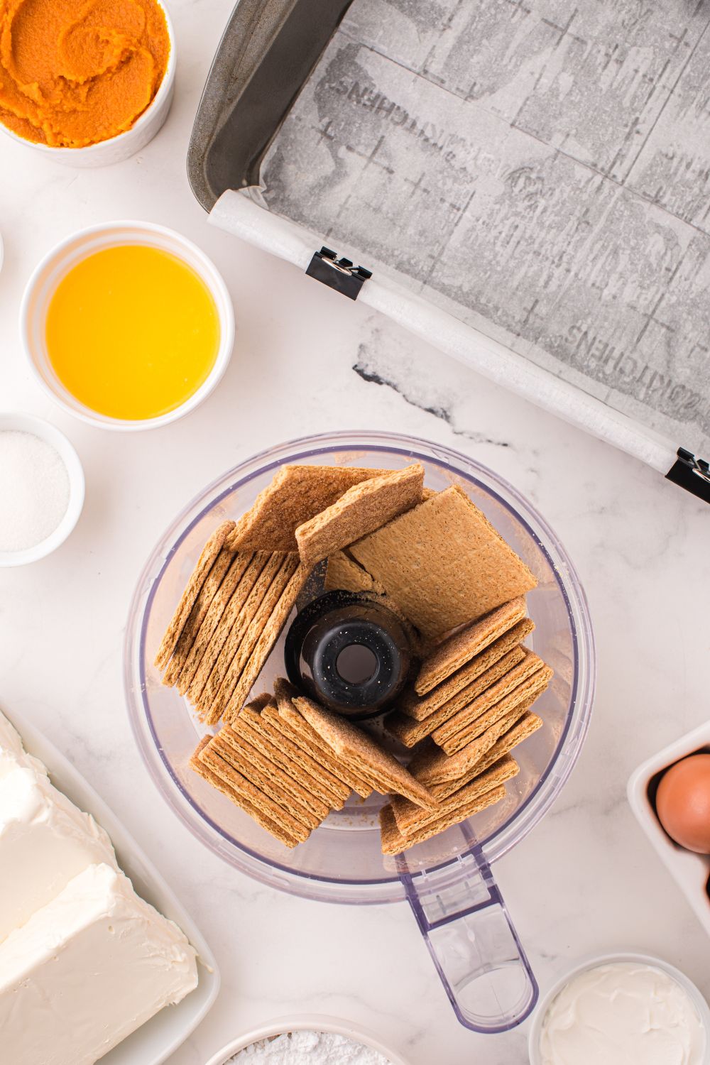 food processor with graham crackers and butter and sugar in small bowls to side for making Swirled Pumpkin Cheesecake Bars