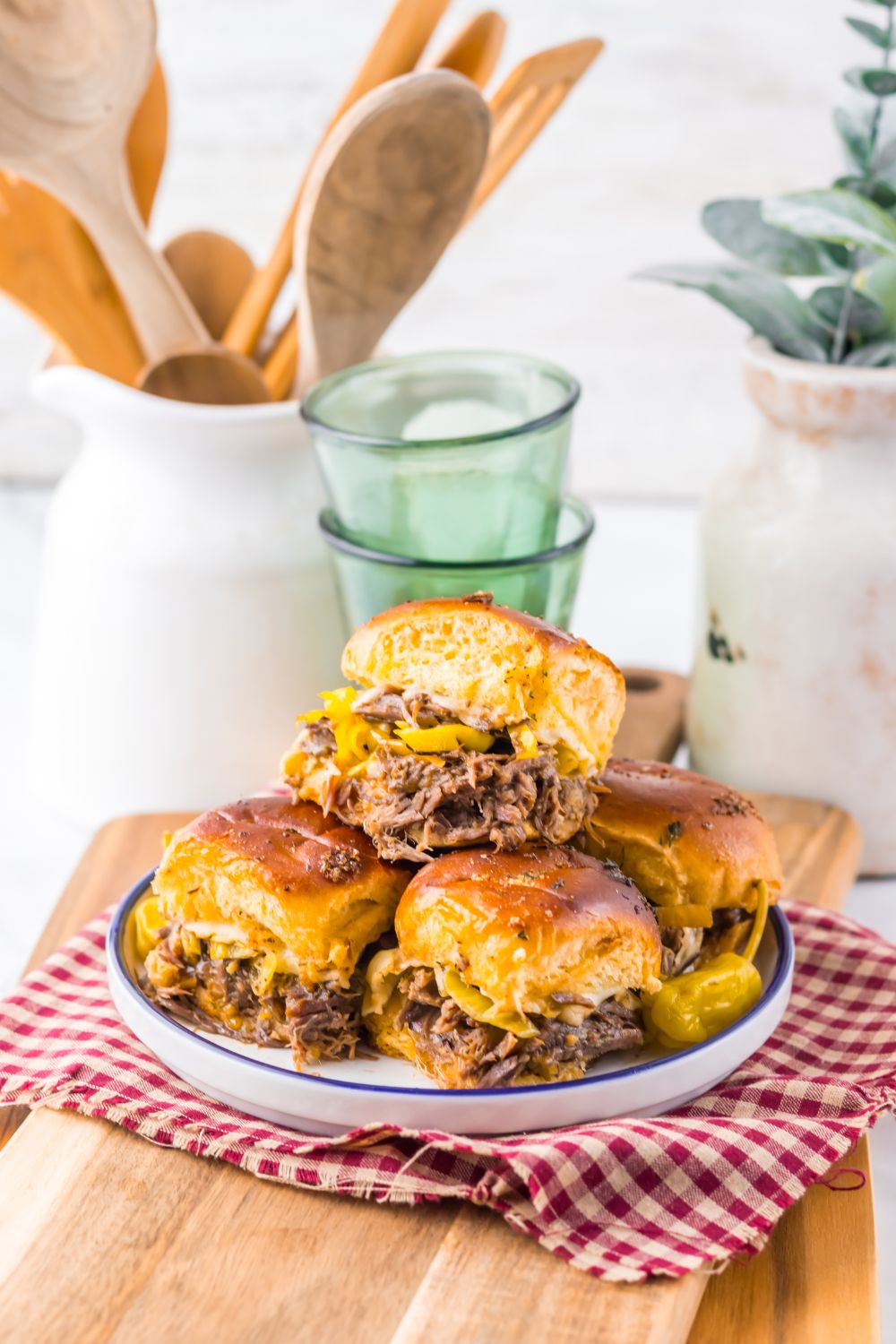 Slow Cooker Mississippi Pot Roast Sliders on a plate with drinks behind it and a jar full of wooden kitchen utensils 