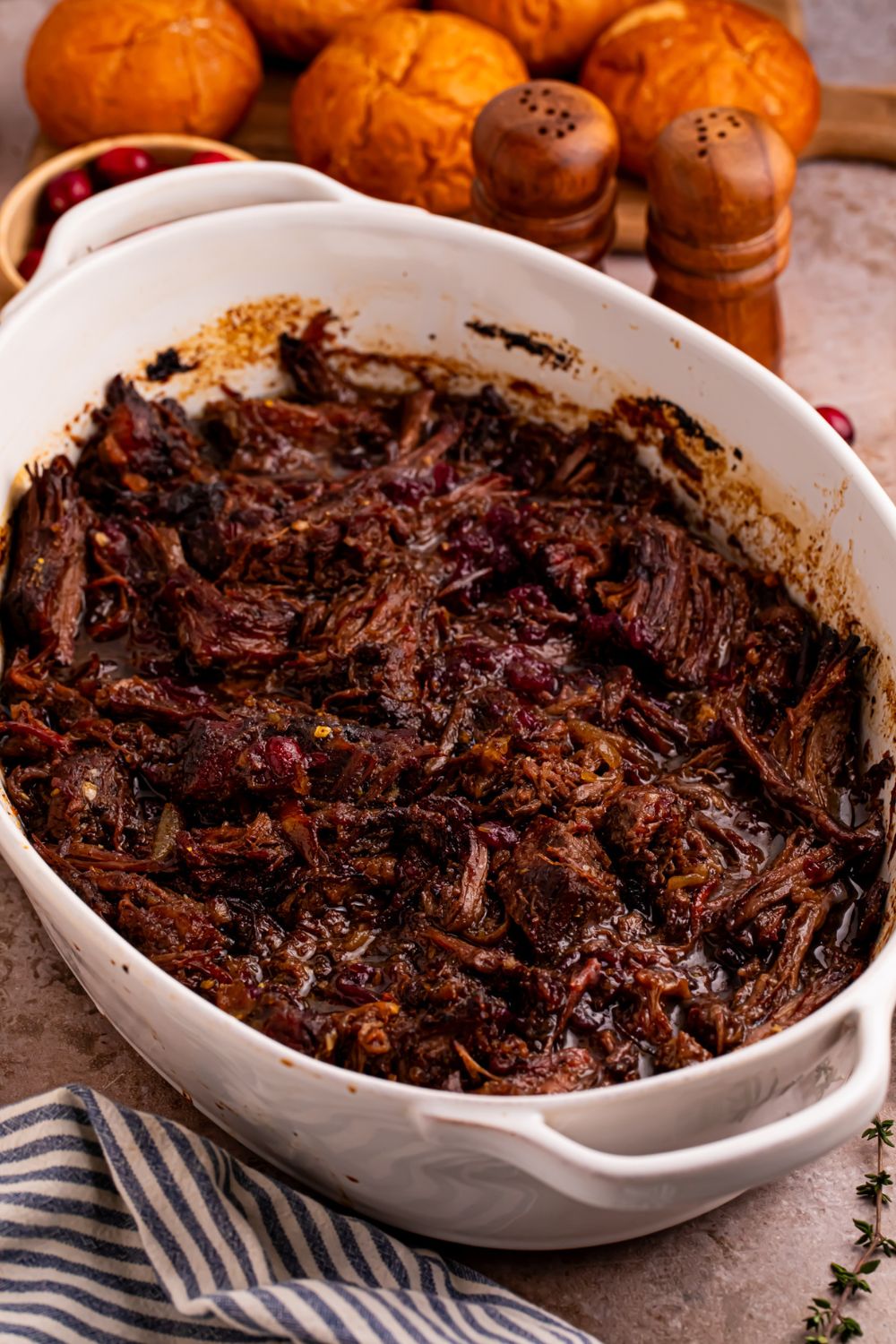 shredded cranberry pot roast in a casserole pan on table with dinner rolls behind it on table