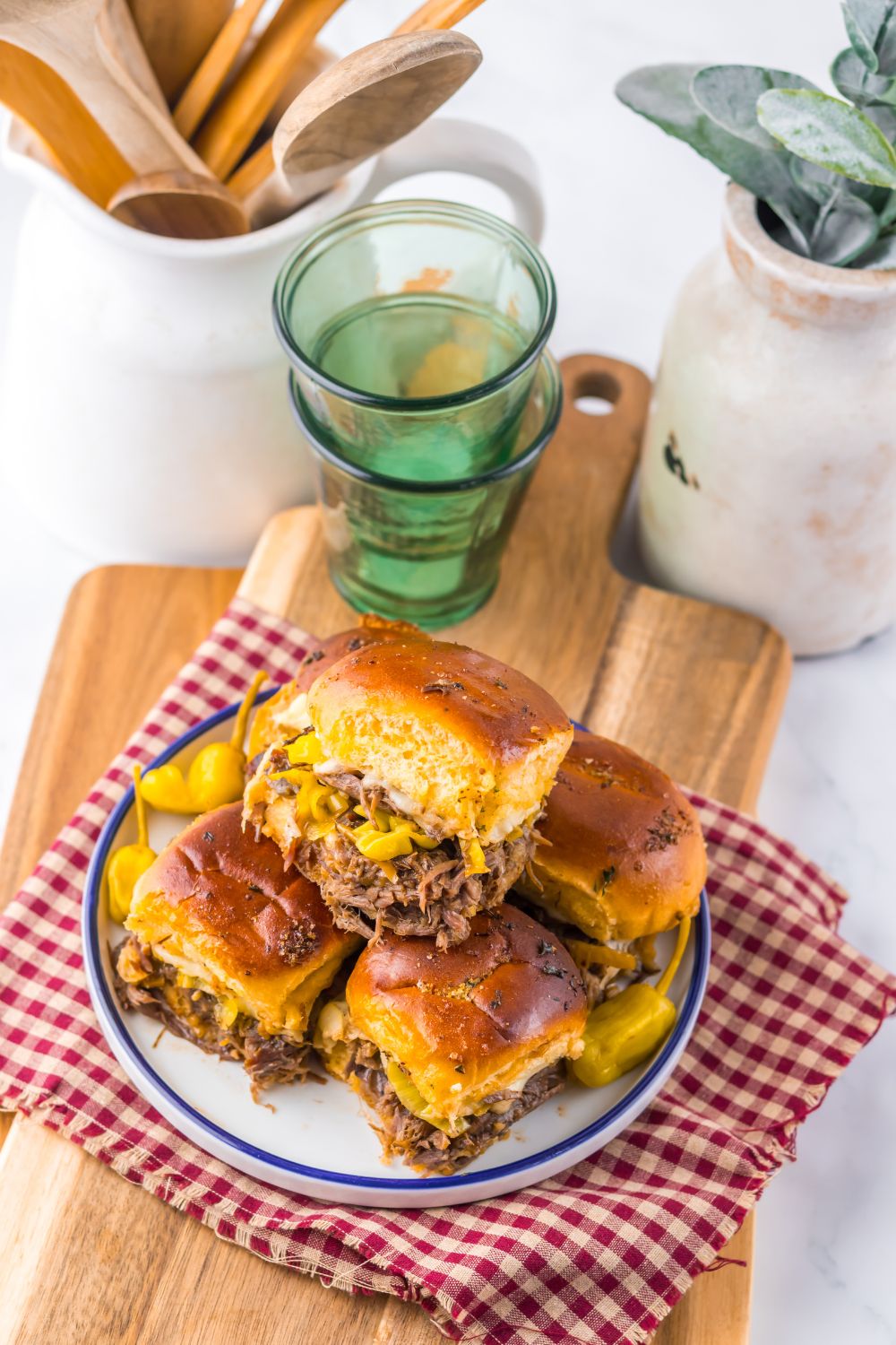 Slow Cooker Mississippi Pot Roast Sliders on a table setting with glasses, cutting board and drink glasses behind it on table 