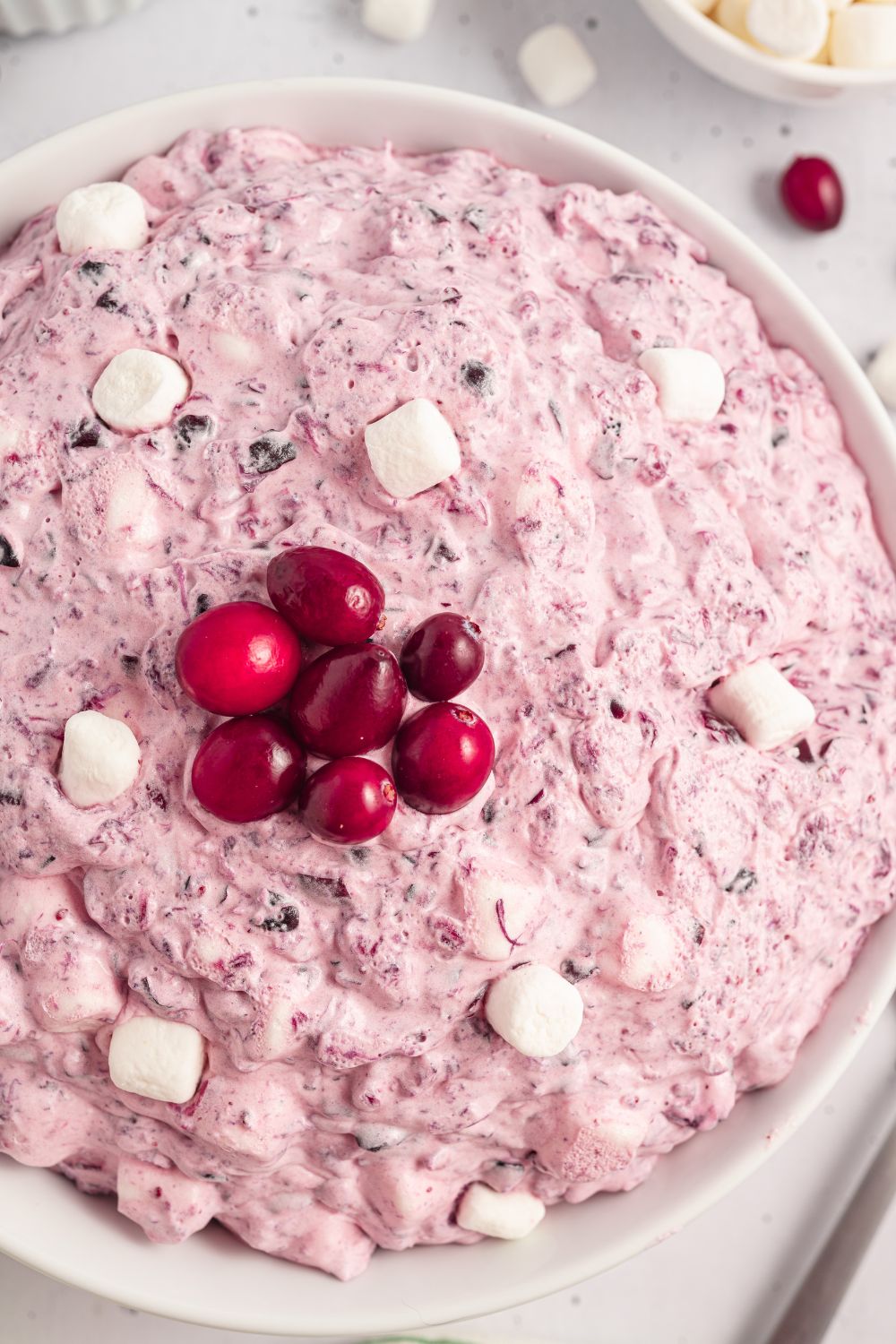Cranberry Fluff Salad in a white bowl with mini marshmallows on top and fresh cranberries. Then in another bowl is mini marshmallows on marble counter 